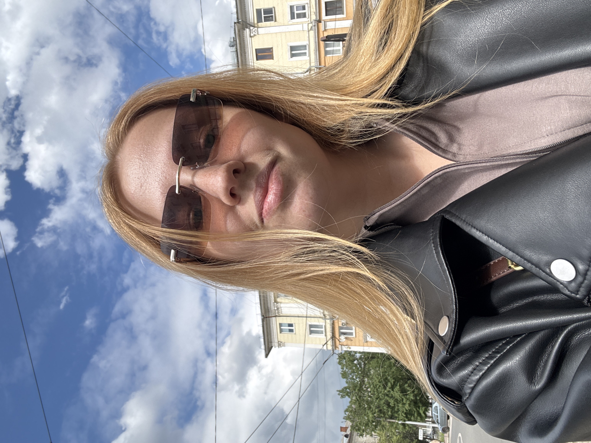 A selfie image shows a person with long blonde hair wearing sunglasses and a black leather jacket. They are outdoors in Заволжье, Russia, on a street with buildings in the background. The sky is blue with clouds.