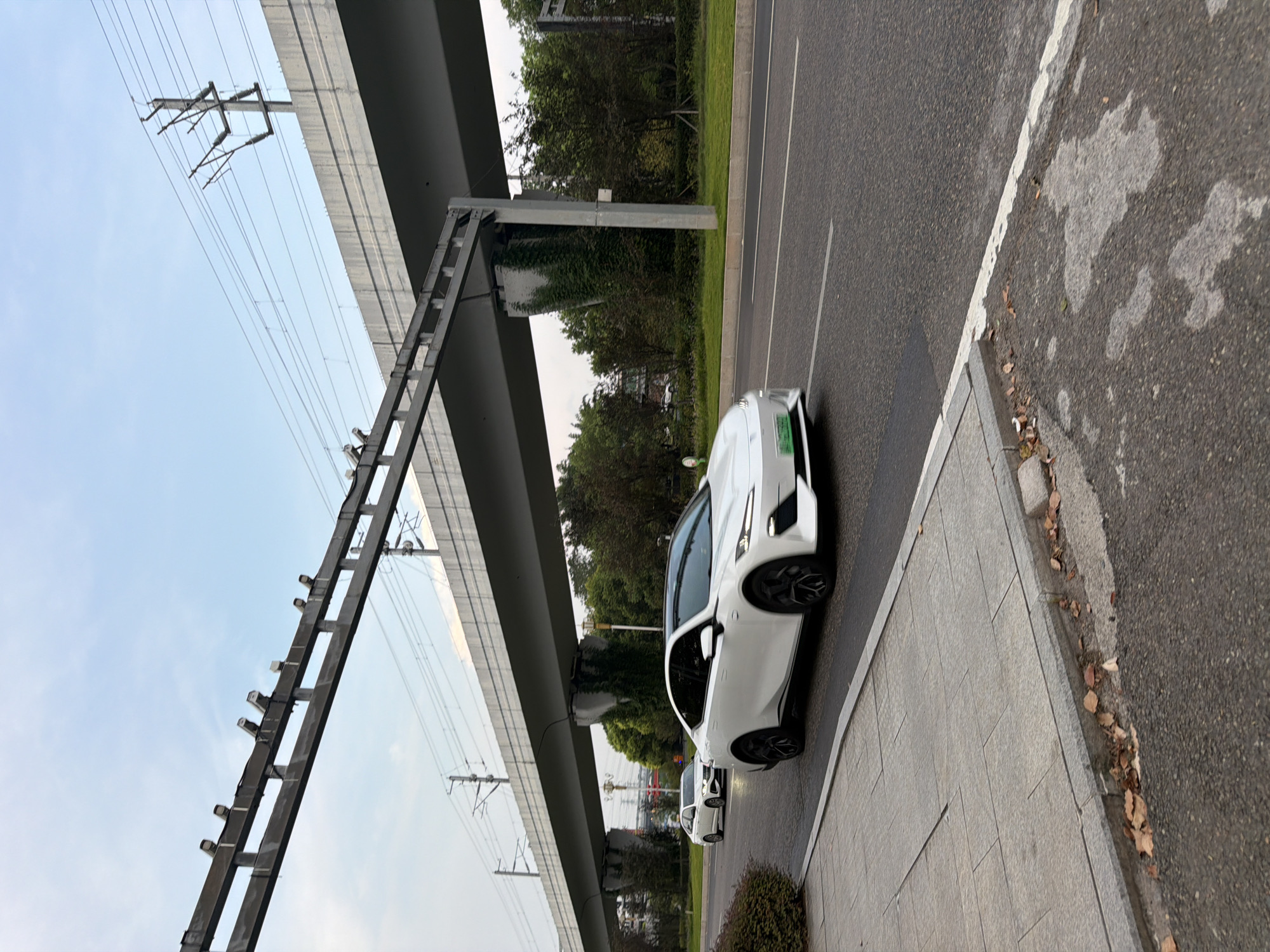 A white electric car, identified by a green license plate, is traveling on a paved road in Jiangjin, China. The car is the primary subject, positioned in the foreground and midground, with its front right side facing the viewer. Its headlights are on, and it appears to be moving forward. Another white car is visible in the background, further down the road.
The scene is characterized by an elevated structure, possibly a highway overpass or a railway viaduct, spanning across the image. This structure is supported by concrete pillars, some of which are covered in ivy. Above the elevated structure, power lines and utility poles are visible against a cloudy sky.
To the left of the road is a paved sidewalk made of large, rectangular grey stones, bordered by a curb. Some dry leaves are scattered on the sidewalk and the edge of the road. Beyond the sidewalk, there is a dense cluster of green bushes. The background is filled with trees, suggesting an urban or suburban setting with some greenery. The overall environment appears to be an outdoor, daytime scene.