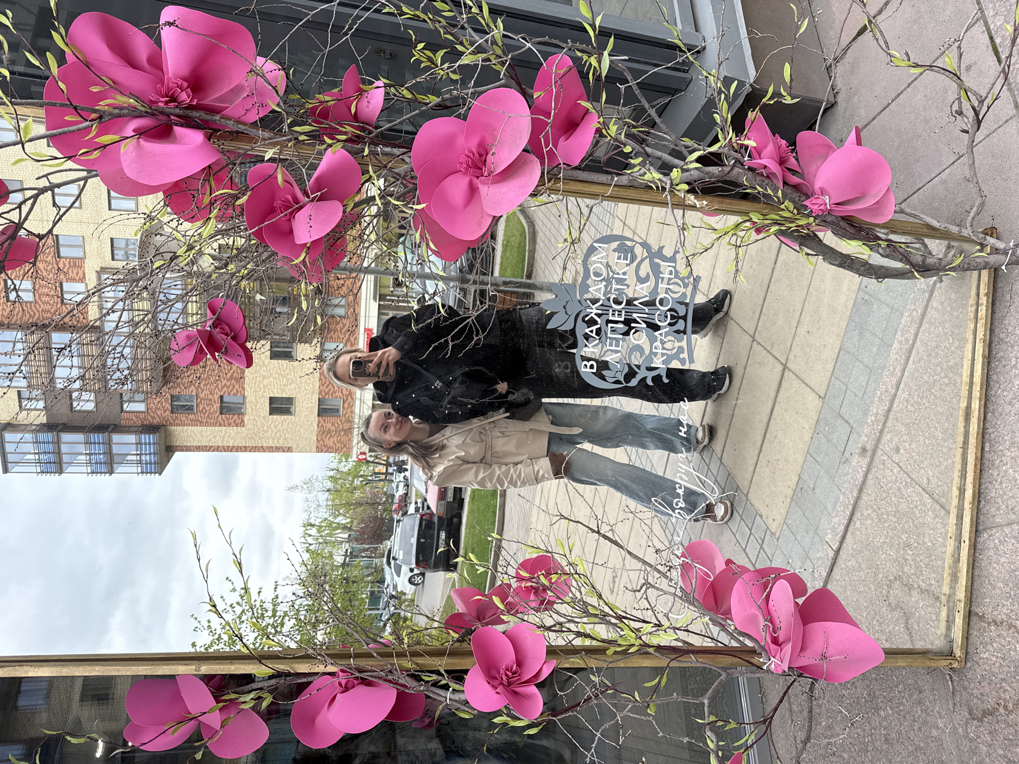 In Krasnoyarsk, Russia, a framed mirror decorated with faux pink flowers and branches reflects a couple taking a selfie. The man wears a black jacket and dark pants, while the woman is in a beige jacket and light blue jeans. The reflection shows a street scene with parked cars, trees, and a multi-story brick building under a cloudy sky. A decorative plaque with Russian text is visible within the mirror's reflection. The mirror sits on a tiled sidewalk.