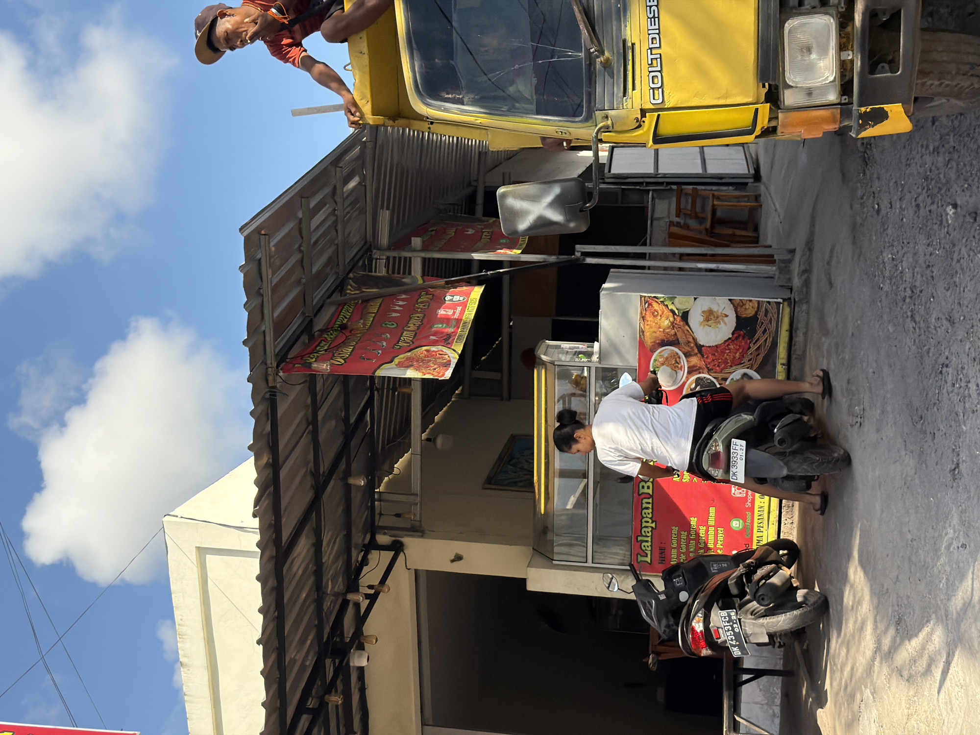 This vibrant daytime scene, set in Kuta Selatan, Indonesia, is captured under a bright blue sky with scattered white clouds. On the left side of the frame, a yellow "COLT DIESEL" truck is parked, and a man wearing a hat is leaning out of its cab window, looking down towards the street level. To the right, a bustling food stall is visible, featuring glass display cases and multiple red banners. One prominent banner advertises "Lalapan B" and lists menu items such as "Ayam Goreng," "Nila Goreng," and "Ayam Geprek," also indicating availability through "Shopee Food" and "Grab Food" delivery services, with a phone number "0777 5025" visible. A person in a white t-shirt and dark shorts stands by the food stall, seemingly attending to it, next to a black motorcycle with the license plate "DK 3933 FF," valid until 01.27. Another black motorcycle, bearing the license plate "DK 4353FCB" valid until 03.26, is parked closer to the foreground. The image portrays a typical outdoor commercial area with daily activities involving local transport and food vending. The visible Indonesian text on the banners and the "DK" prefix on the license plates serve as clear location cues. The clear skies suggest good weather, likely morning or early afternoon.