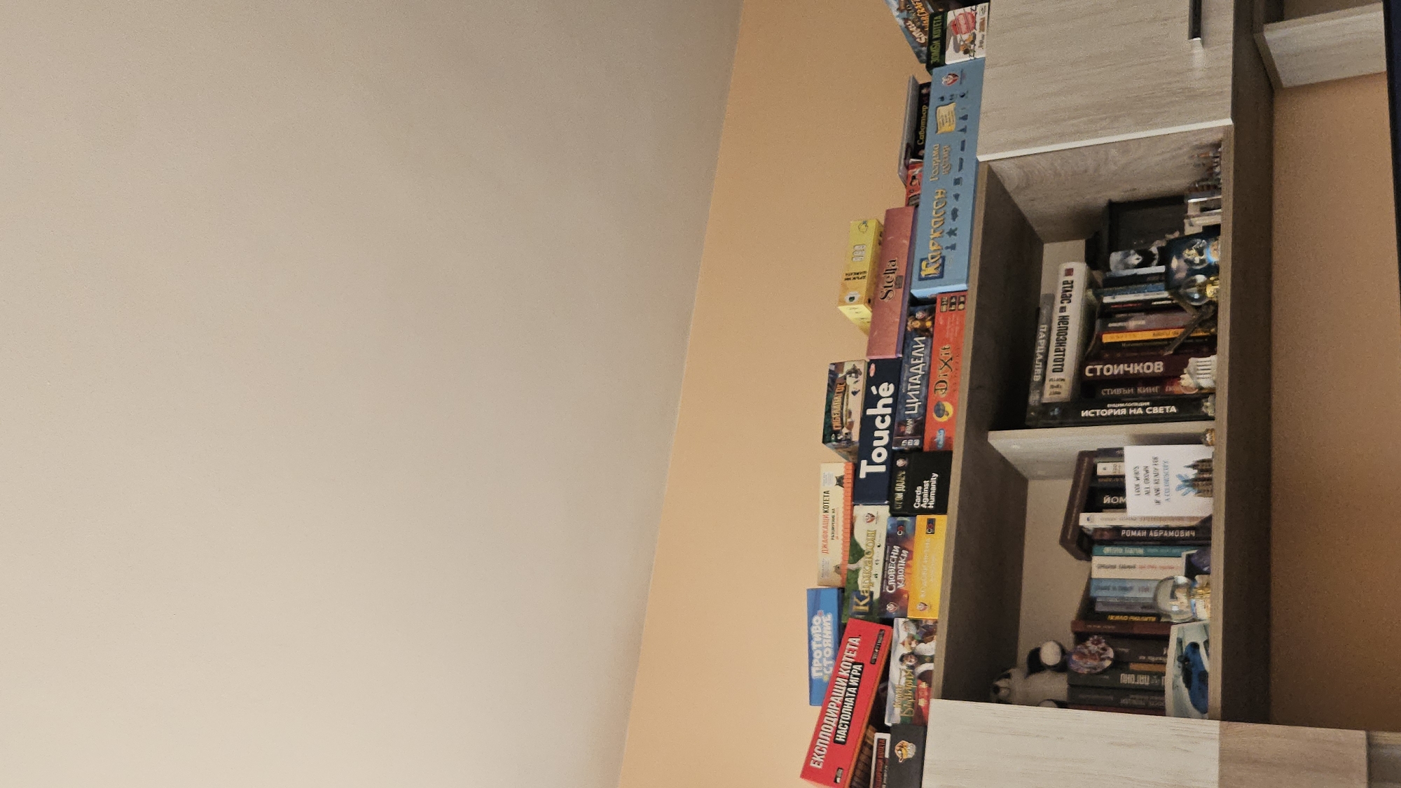 A cabinet with shelves is filled with board game boxes, books, and other items. The board games include "Stella", "Carcassonne", "Exploding Kittens", "Cards Against Humanity", "Dixit", and other games written in Bulgarian. The books include what appears to be a biography of football player Hristo Stoichkov. The cabinet is likely located in Plovdiv, Bulgaria, based on the text on the books and games.