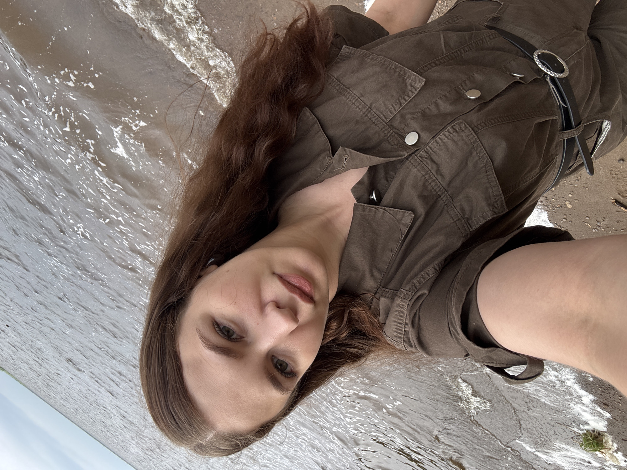 This is a vertically oriented, close-up, self-portrait of a young woman. She has long, wavy, dark brown hair that falls around her face and shoulders. Her skin is fair, and her eyes are a light green. She is wearing a dark olive green, button-up jumpsuit with prominent chest pockets and a black leather belt with a circular silver buckle. The jumpsuit appears to be made of a textured fabric.
The woman is positioned in what seems to be an outdoor setting. Behind her, the ground appears to be sandy or muddy, with some small twigs and debris. In front of her, there is water with visible ripples and reflections, suggesting it might be a lake, river, or shallow sea. The lighting indicates it is daytime, and the overall atmosphere suggests a casual, possibly relaxed or contemplative mood. The specific location cues are minimal, but the provided context places this scene in Chaikovsky, Russia. There is no visible text in the image. The woman's gaze is directed towards the camera, and her expression is neutral to slightly melancholic.