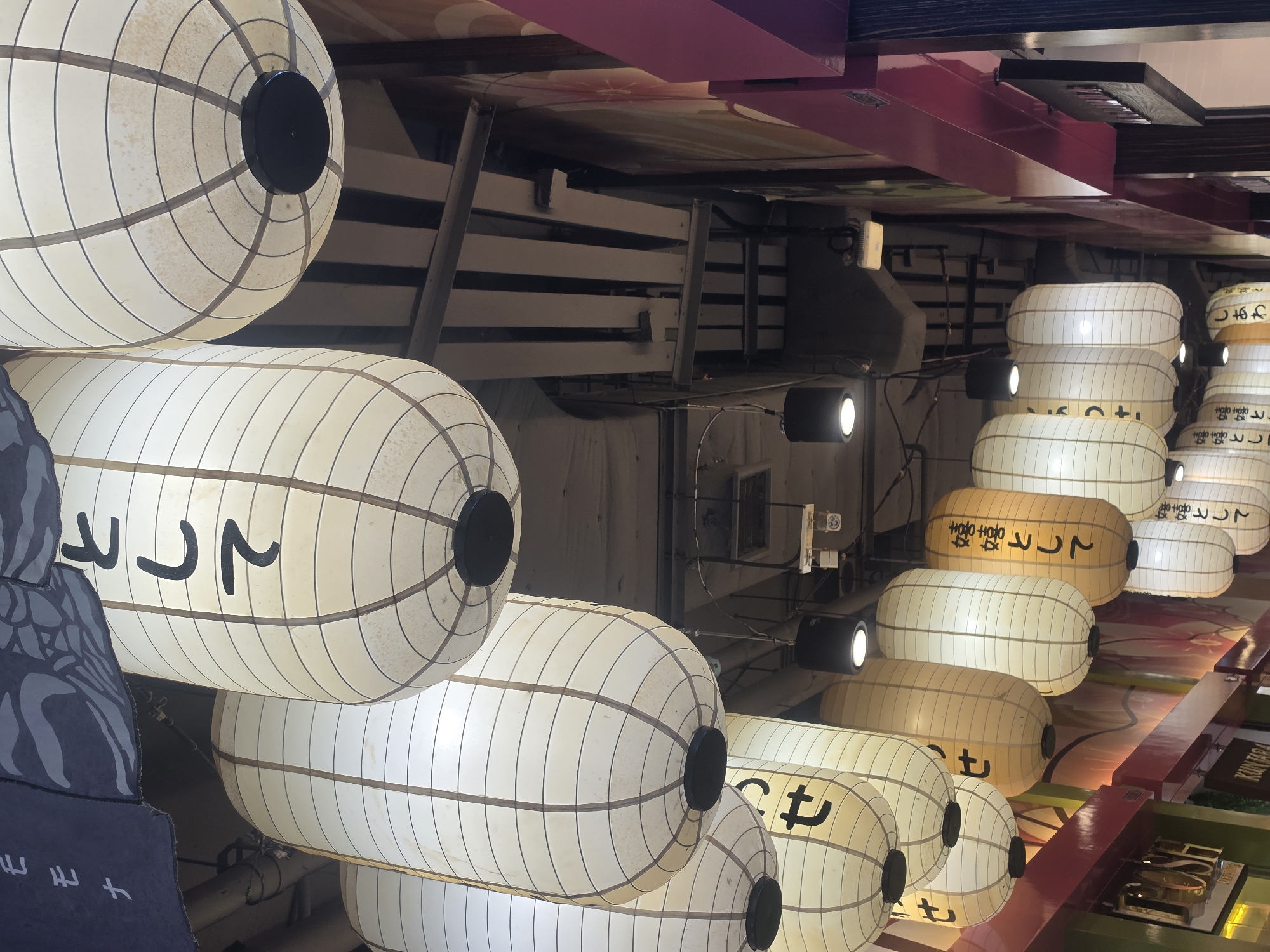 The image displays an indoor commercial space, likely a Japanese-themed restaurant or bar in Bangkok, Thailand, captured with a 90-degree clockwise rotation. The prominent features are numerous cylindrical paper lanterns, illuminated from within and suspended in rows.
These lanterns are mostly off-white or cream, with some appearing in a yellowish-orange shade. Many are adorned with black Japanese characters, including phrases like "として" (as/in the capacity of), "せ", "嬉嬉として" (gleefully/joyfully), "しあわ" (part of "happiness"), "嬉嬉と", "嬉嬉とし", and "嬉として". The paper construction with a visible grid pattern is evident on the lanterns.
Interspersed with the lanterns are modern spotlights, casting additional illumination. The background reveals structural elements such as dark wooden beams and possibly metal frames, along with colorful ceiling panels, some featuring pink and other patterns. On the far left, a dark, irregularly shaped decorative piece with white or light grey Japanese text is partially visible.
The scene is devoid of people, focusing on the decor. The artificial lighting suggests an evening or nighttime setting, creating a warm and inviting atmosphere. On the far right, a partial sign is visible, reading "KUN TARA" and "FOSH", likely indicating the establishment's name.
