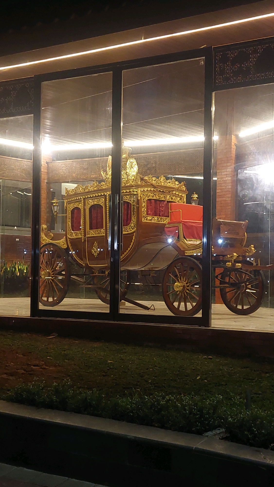 A golden and brown ornate carriage is displayed inside a glass enclosure at night. The carriage has large wooden wheels, intricate gold detailing, and red cushioned seats. The enclosure has a modern architectural design with wooden paneling on the ceiling and brick walls visible in the background. The exterior includes a grassy area with some sparse greenery. The scene appears to be in Cibinong, Indonesia, at an exhibition or museum showcasing historical artifacts.