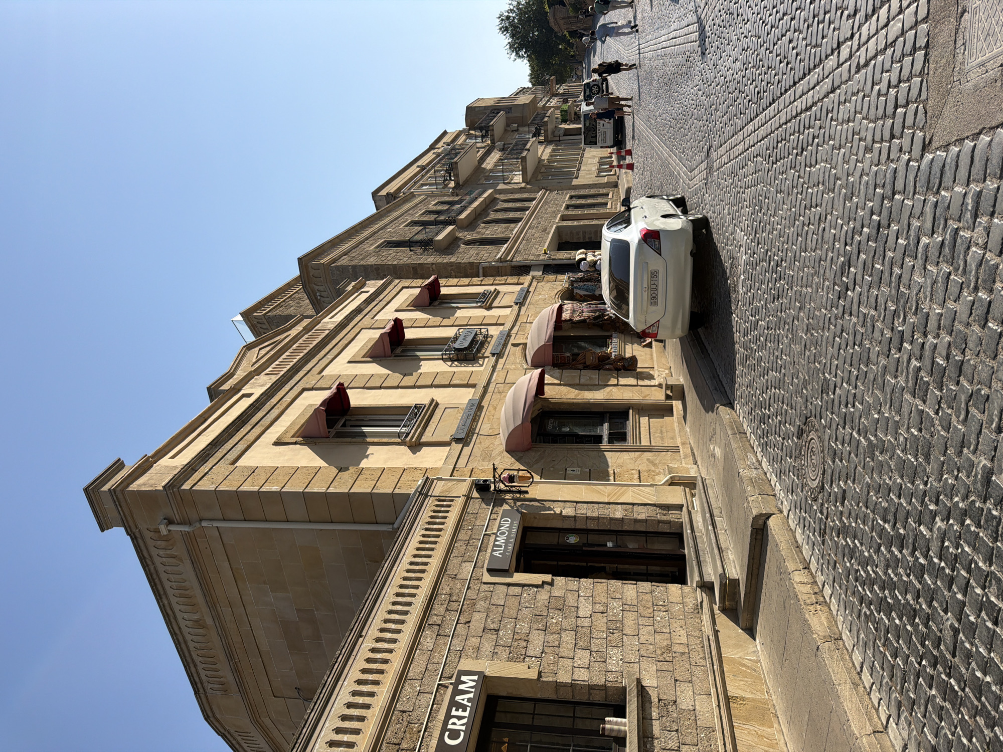 A cobblestone street curves uphill in Sabail Raion, Azerbaijan, lined with traditional stone buildings under a clear blue sky. A white sedan with license plate 90-UJ-155 is parked on the right side of the street. People are walking uphill in the distance. Several businesses are visible, including "CREAM," "ALMOND - CAKE & BAKERY," and a "COFFEE BAR." The architecture features stone facades, arched windows, and awnings. The street is paved with dark, uneven cobblestones.