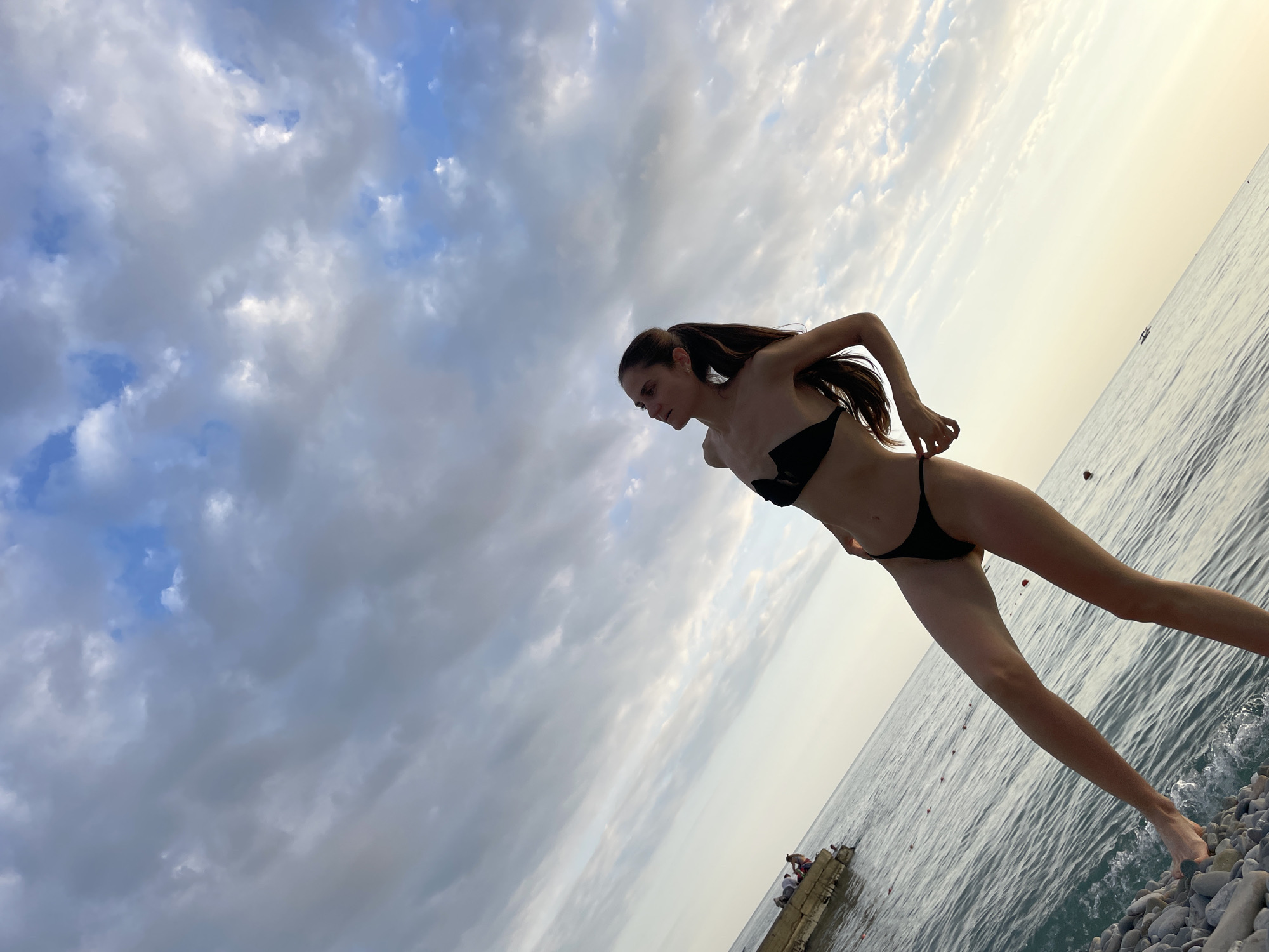 A full-body, low-angle shot captures a slender woman wearing a black bikini against a backdrop of the sky and the sea. She stands on a pebble beach, her left leg extended forward, and her right leg bent at the knee. The woman is facing to her right, with her head tilted back slightly and her gaze directed upwards. Her right hand is extended, with fingers touching the string of her bikini bottoms. Her long, dark hair is pulled back into a ponytail.
In the background, the sea stretches to the horizon, meeting a pale yellow sky with scattered clouds. To the left of the frame, a concrete structure, possibly a pier or breakwater, juts into the water. Several people are seated or standing on this structure. The water is calm, with gentle waves lapping at the shore. The scene appears to be set in Sochi, Russia, given the characteristic pebble beaches and the warm, hazy light of a coastal sunset.
