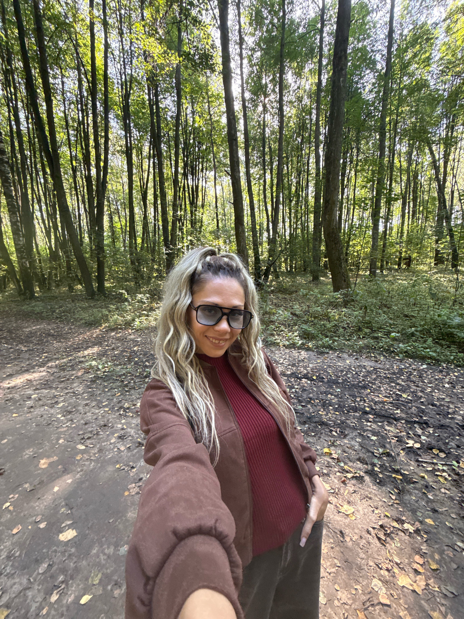 The image captures a woman taking a selfie in a forest setting. She is wearing a brown jacket over a maroon ribbed sweater and dark pants. She has blonde hair styled in a high ponytail and is wearing large, dark sunglasses. The background is a dense forest with tall trees and a path covered in fallen leaves. Sunlight filters through the canopy, creating dappled light on the forest floor. The location is identified as Rusanovo, Russia.