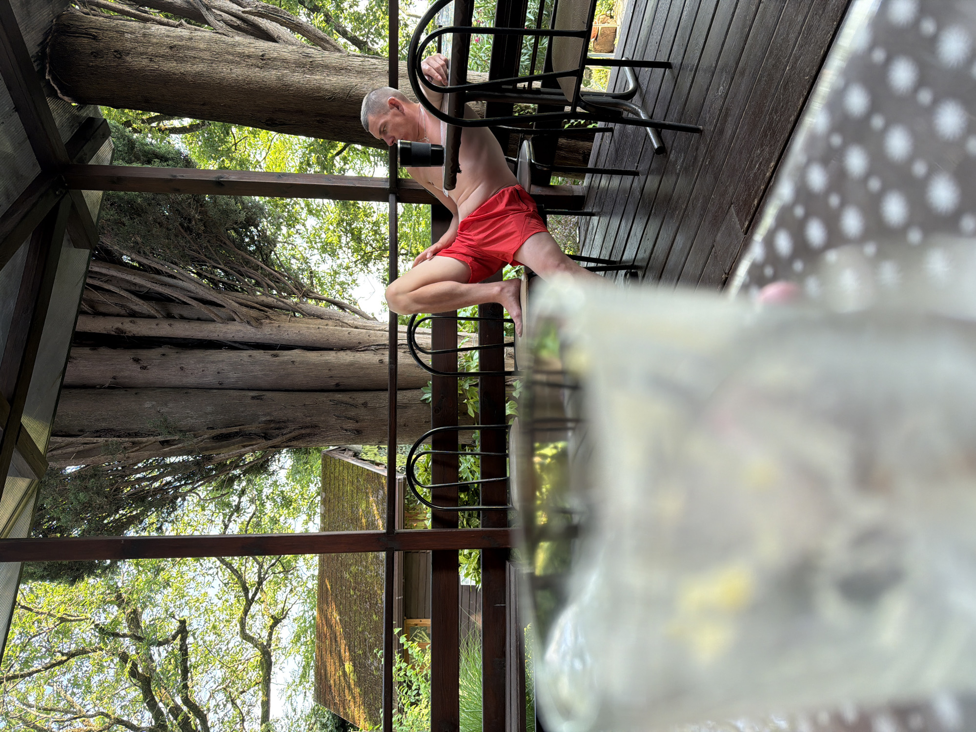 A man is captured in a media file, seated on a chair outdoors, partially undressed. He is wearing red shorts and is bare-chested. The man is looking down towards his lap, with his left hand resting on a black cylindrical object, possibly a camera lens. He appears to be engaged in some activity related to this object. The setting is an outdoor wooden deck, possibly part of a veranda or balcony, surrounded by lush greenery and large trees. The image is taken from a low angle, with a large, blurry glass object in the foreground, obscuring part of the view. The lighting suggests it is daytime, with dappled sunlight filtering through the trees. The overall atmosphere seems relaxed and informal. Sochi, Russia is the given location context.