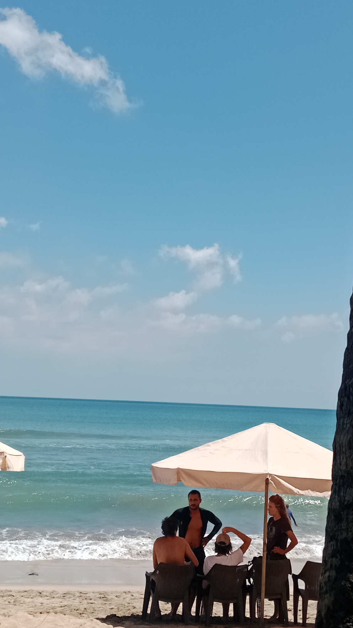 A group of four individuals are gathered under a beige beach umbrella on a sandy shore. Two men are seated in chairs facing away from the camera, while another man stands, wearing a black wetsuit top. A woman stands to the right of the group, facing the standing man. The ocean with gentle waves is visible behind them, and the sky is clear with scattered clouds. The scene appears to be a casual gathering on a beach. The location is identified as Kuta, Indonesia.