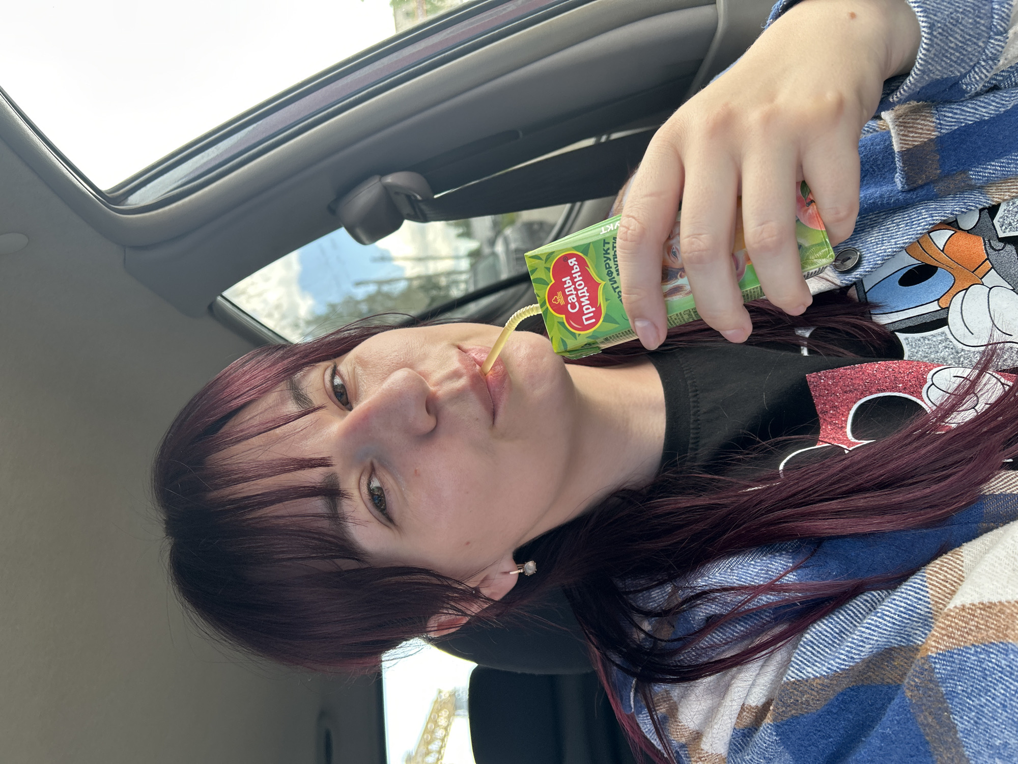 A young woman with long, dark red hair and bangs is sitting in the driver's seat of a car and drinking from a juice box. She is wearing a blue and white plaid shirt over a black t-shirt with a Disney character graphic. The juice box is green and has Russian text and images of fruit. The car interior, including the seatbelt and door panel, is visible. The background shows a cloudy sky and some buildings, suggesting an urban or suburban setting. The image was taken in Chaikovsky, Russia.