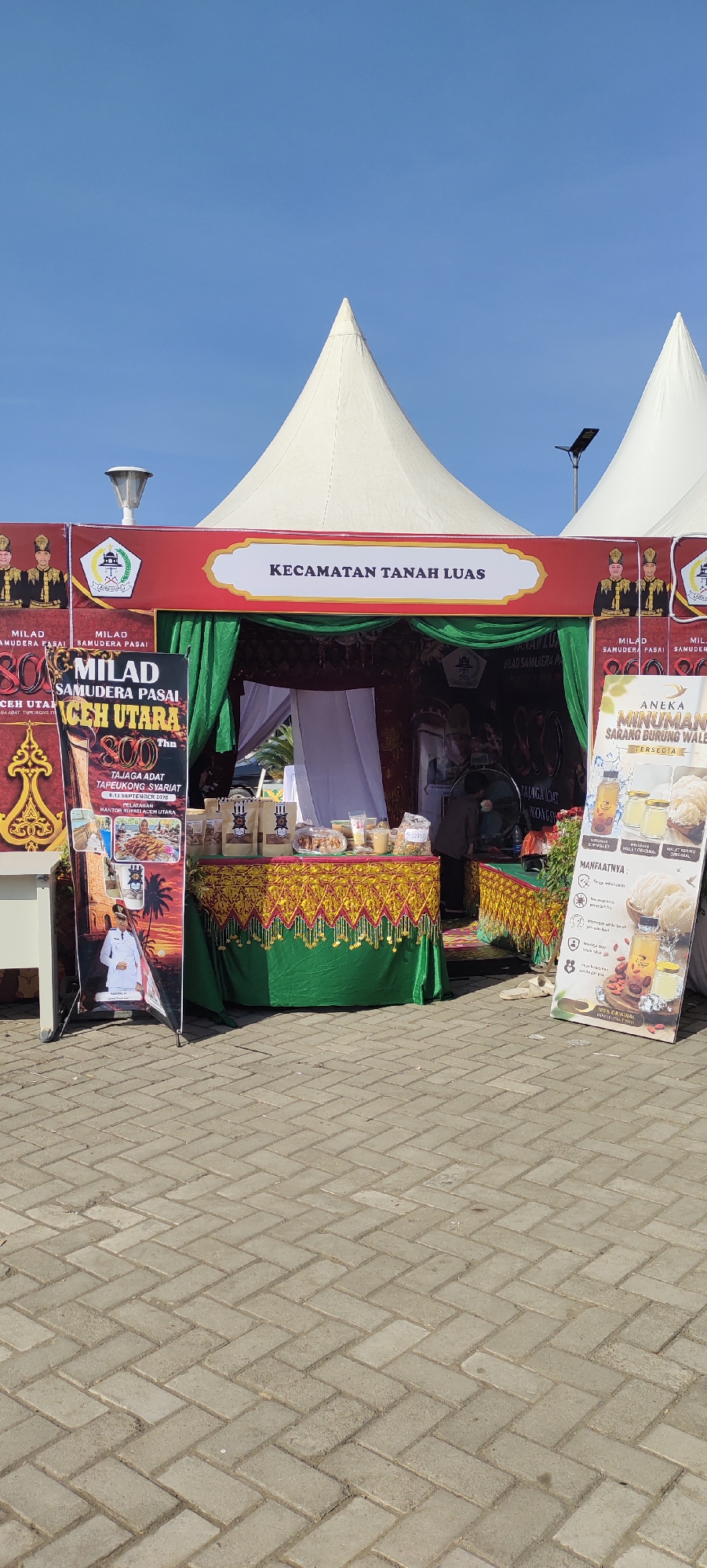 In the city of Meunasahaluedrienlhoksukon Barat, Indonesia, a stall with a red awning and a white banner displaying "KECAMATAN TANAH LUAS" is set up outdoors under a clear blue sky. The stall features a green curtain and a table draped with a gold and green patterned cloth, displaying various products including packaged food items and bottles of liquid. To the left, a large banner shows images of people and text related to "MILAD SAMUDERA PASAI ACEH UTARA 800 THN TAJAGA ADAT TAPEUKONG SYARIAT." To the right, another banner advertises "ANEKA MINUMAN SARANG BURUNG WALET" with information about its benefits. Two white conical tents are visible behind the stall, and the ground is paved with interlocking gray bricks. A person is partially visible inside the stall, behind the table.