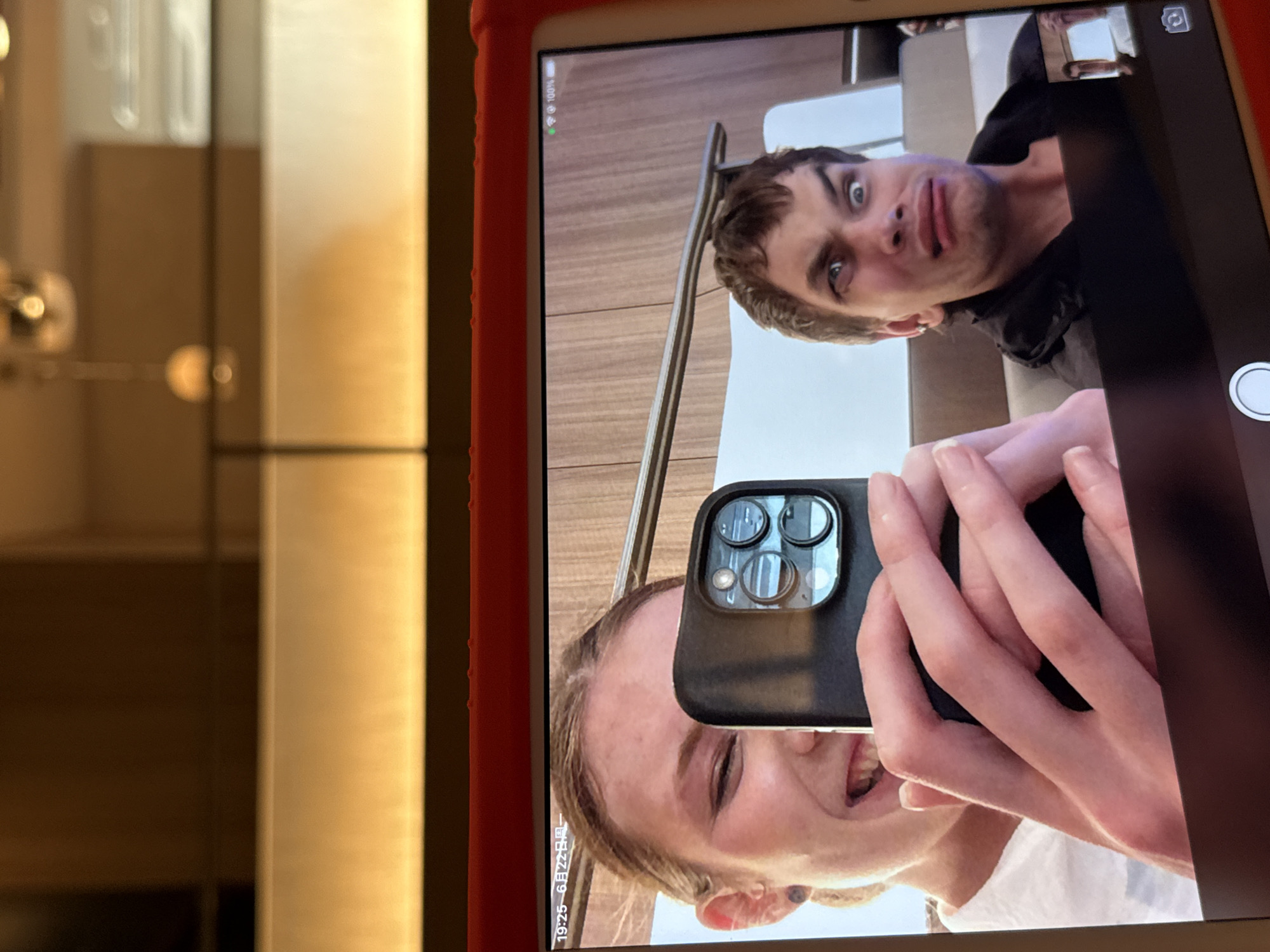 This image shows a tablet screen displaying a video call between two individuals. On the left, a young woman with blonde hair pulled back smiles widely and holds up a black smartphone, partially obscuring her face. Her right hand is visible, holding the phone which has a triple-lens camera system. On the right side of the screen, a young man with brown hair makes a distorted facial expression, his eyes wide and his mouth puckered. He is wearing a black jacket and appears to be sitting on a seat. A small thumbnail image of the woman is visible in the bottom right corner of the man's frame. The tablet screen shows a date and time stamp at the top left that reads "19:25 6月22日周一," indicating it is 7:25 PM on Monday, June 22nd. The battery level indicator at the top right shows 100%. The background on the tablet screen appears to be a wooden-paneled wall. The tablet itself is housed in a red protective case. The background of the original image shows a blurred indoor setting with warm lighting. While the image contains a date and time which could potentially be associated with Shanghai, China, there is no visual information within the image itself to confirm this location.