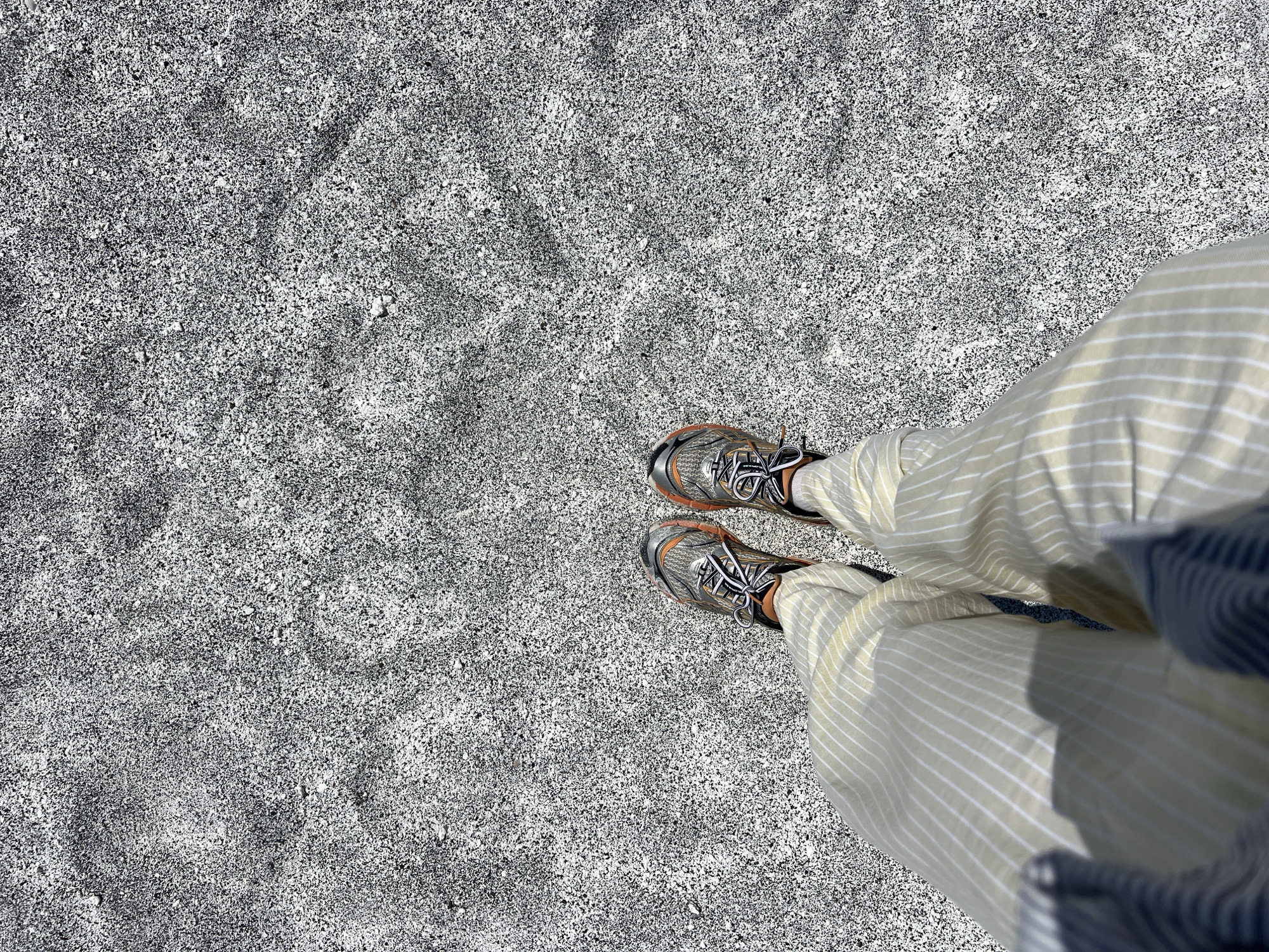 A person's feet, clad in grey and orange athletic shoes and light yellow pants with thin white vertical stripes, are positioned in the lower center of the frame, standing on a surface of dark grey and white pebbles. The pebbled ground stretches across the majority of the image, with a faint heart-shaped indentation visible in the upper right quadrant. The scene is outdoors, with sunlight casting a shadow from the person's legs and left arm onto the pebbled ground. The image's composition suggests a casual, possibly recreational setting, with the pebbles indicating a beach, a dry riverbed, or a similar natural ground cover. The specific location within Yeşilova, Turkey, is not identifiable from the visual information present in the image.