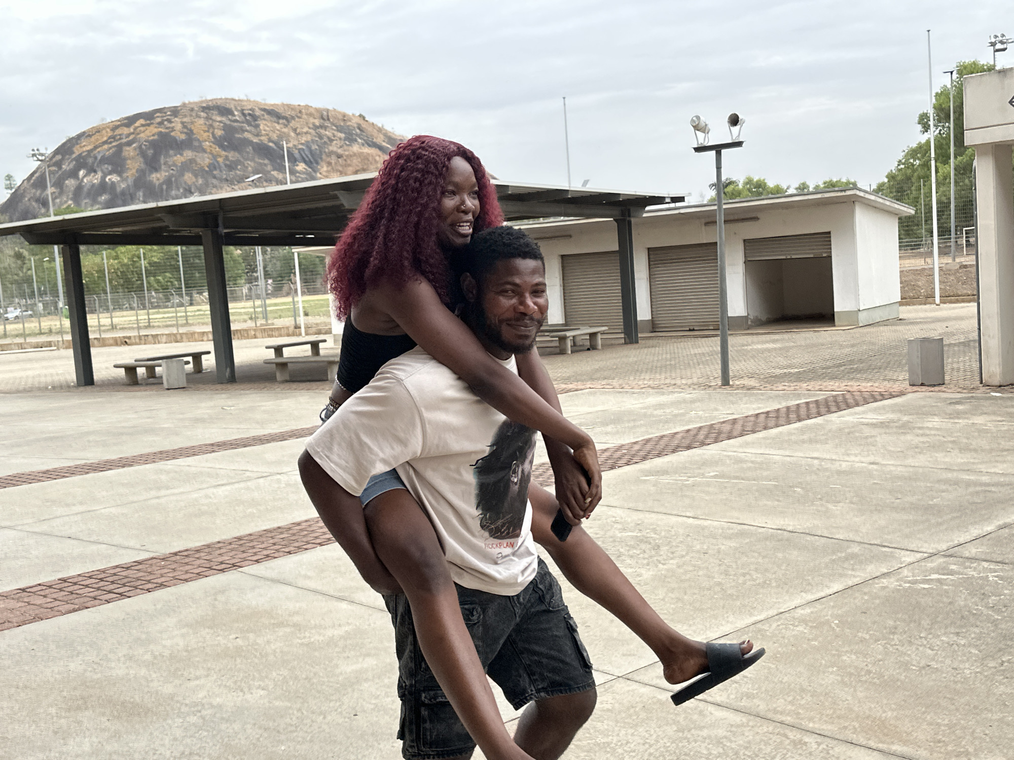 Two individuals are the focus of the photograph. The man is giving the woman a piggyback ride. The woman has long, dark red, wavy hair and is wearing a black tank top. The man is wearing a light-colored t-shirt and dark shorts. They are both smiling. The man is wearing a black sandal on one foot. The photograph appears to have been taken in a public square or park area. A large, rounded hill or mountain is in the background. There are benches and a covered structure, as well as a white building with closed roller doors and tall light poles. The surface they stand on is paved with large, light-colored concrete slabs with narrow bands of red brick. The location is Unknown, Unknown.
