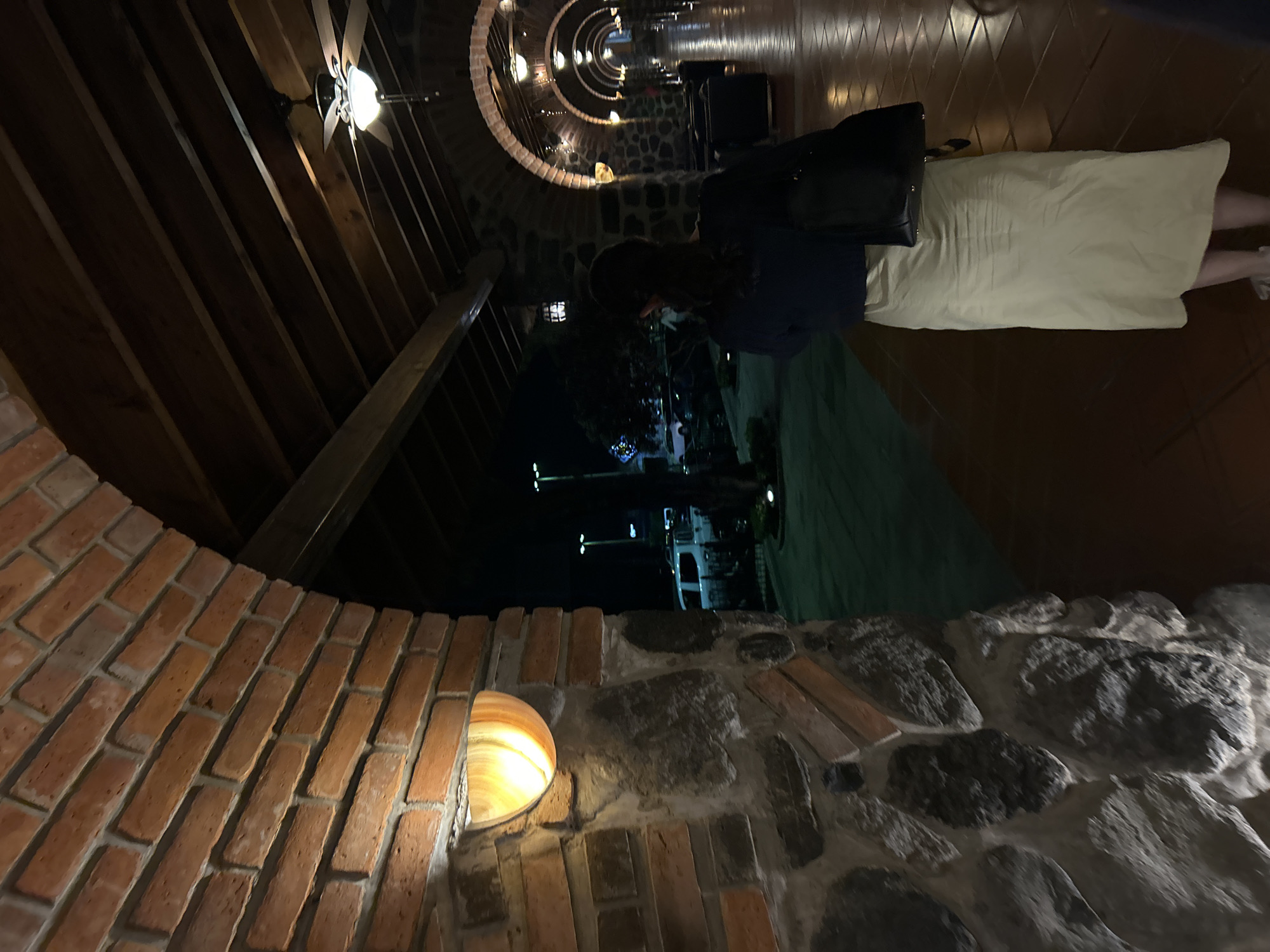 A sideways, canted, medium shot of a brick and rock archway at the Hotel Puerta del Sol in Santa Ana Yancuitlalpan (Chilhuacán), Mexico. A hallway receding into the distance can be seen through the archway. A woman is walking in the hallway with a black handbag. Her skirt is light tan, and she wears a dark top. Her dark hair is shoulder-length. A ceiling fan is visible toward the top left of the image. Outdoor light can be seen through another archway beyond the woman.