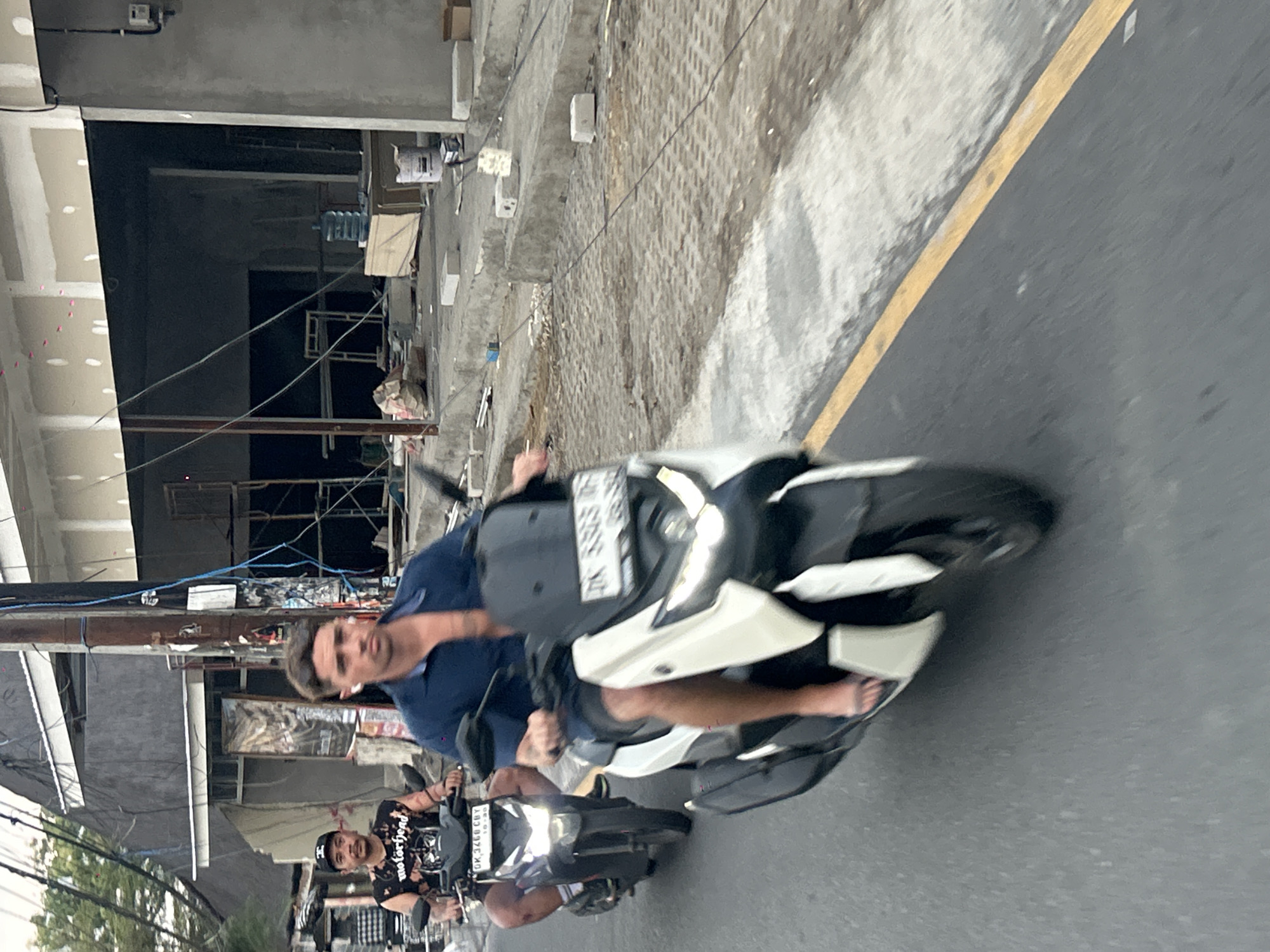 In North Kuta, Indonesia, two men are riding motorcycles on a street under construction. The man in the foreground is wearing a dark blue unbuttoned shirt and shorts, with white earbuds in his ears. He is riding a white and black scooter with a license plate that reads "DK 5393 UP". The man behind him is wearing a black t-shirt with "Motorhead" printed on it, a black baseball cap, and is riding a black motorcycle with a license plate that reads "DK 3466 CBY". Construction materials, scaffolding, and electrical wires are visible in the background, indicating ongoing development. The road surface is paved with some areas of gravel and pavers.