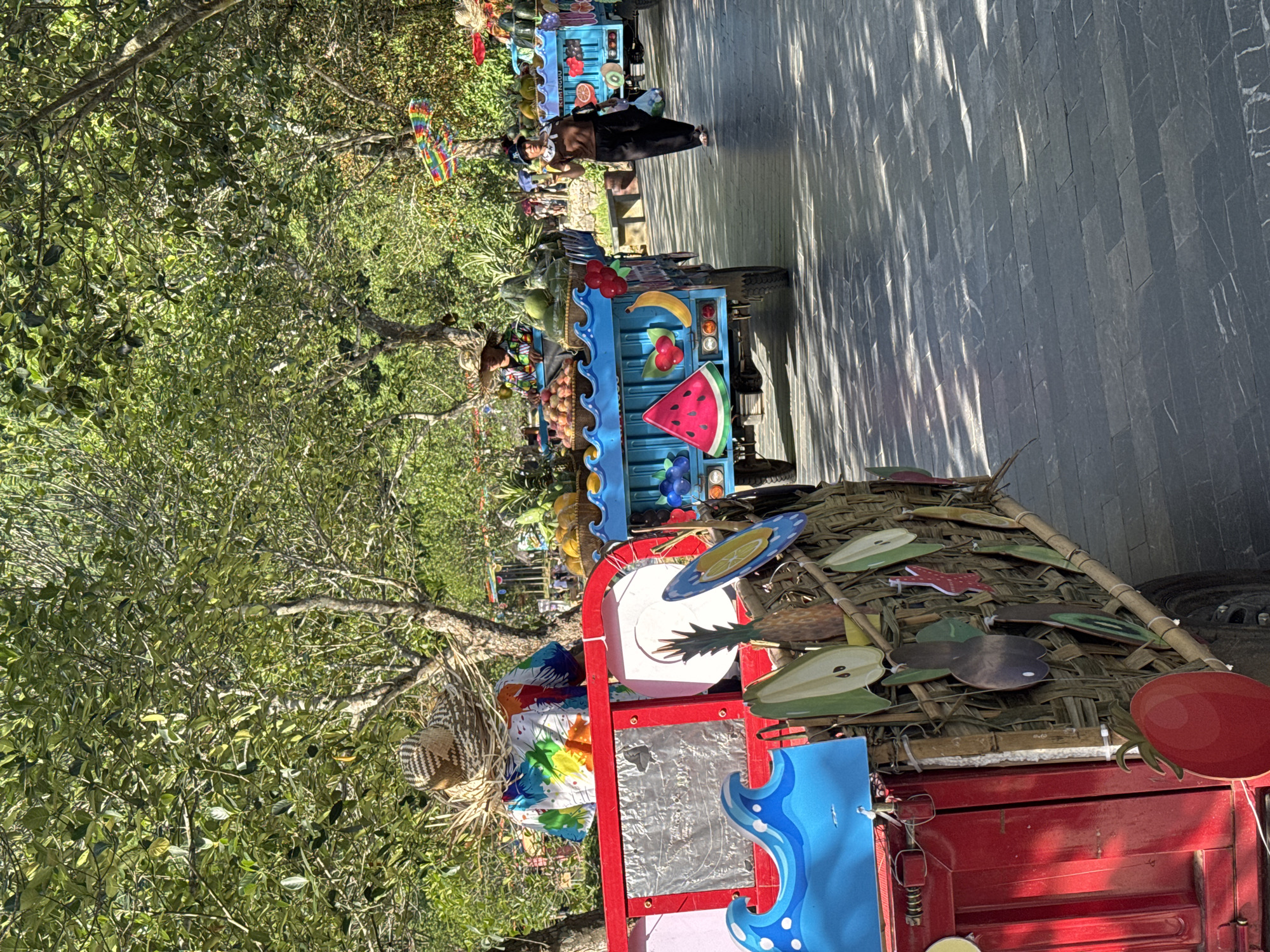 In Hoa Lư Ward, Vietnam, a sun-dappled pathway lined with lush green trees is the setting for a vibrant street scene. Two fruit carts, decorated with colorful cutouts of fruits and wave patterns, are the primary focus. The cart in the foreground, painted red, features a woven bamboo exterior adorned with various fruit shapes. Behind it, a blue cart is laden with an assortment of fruits, including bananas, cherries, and what appears to be a watermelon slice.
A woman in a brown top and black pants walks away from the blue cart, carrying a white plastic bag and a small clear bottle. In the distance, another fruit cart, also blue, is visible, and beyond that, a woman wearing a colorful shirt and a straw hat stands behind a cart filled with produce. The overall atmosphere is lively and festive, with kites in rainbow colors hanging from trees in the background, suggesting a celebratory event or a busy market day. The pathway is paved with dark, rectangular stones, and dappled sunlight creates patterns of light and shadow.