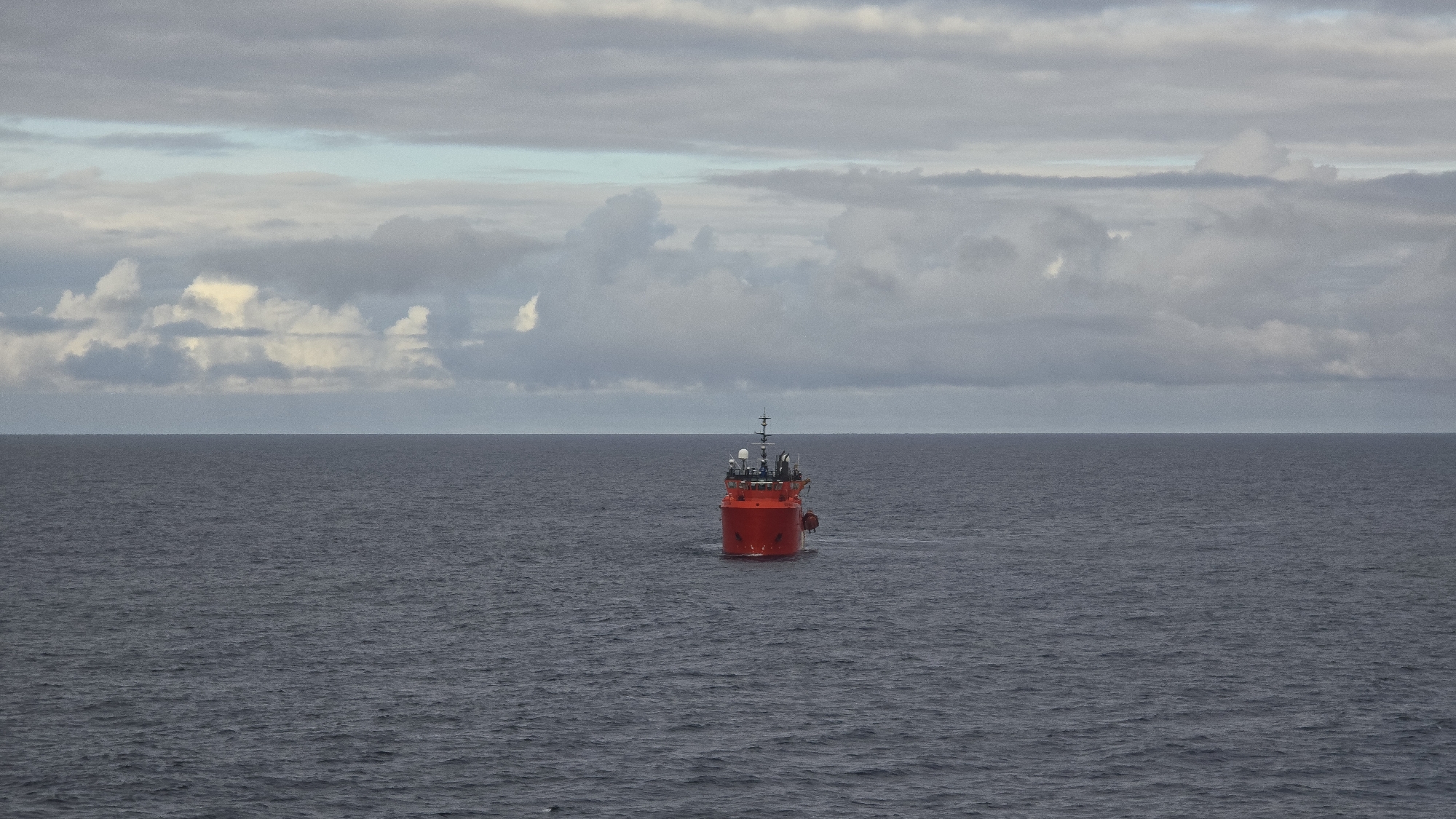 A red ship is in the center of the frame on a calm body of water. The sky is overcast with grey clouds, obscuring any location.