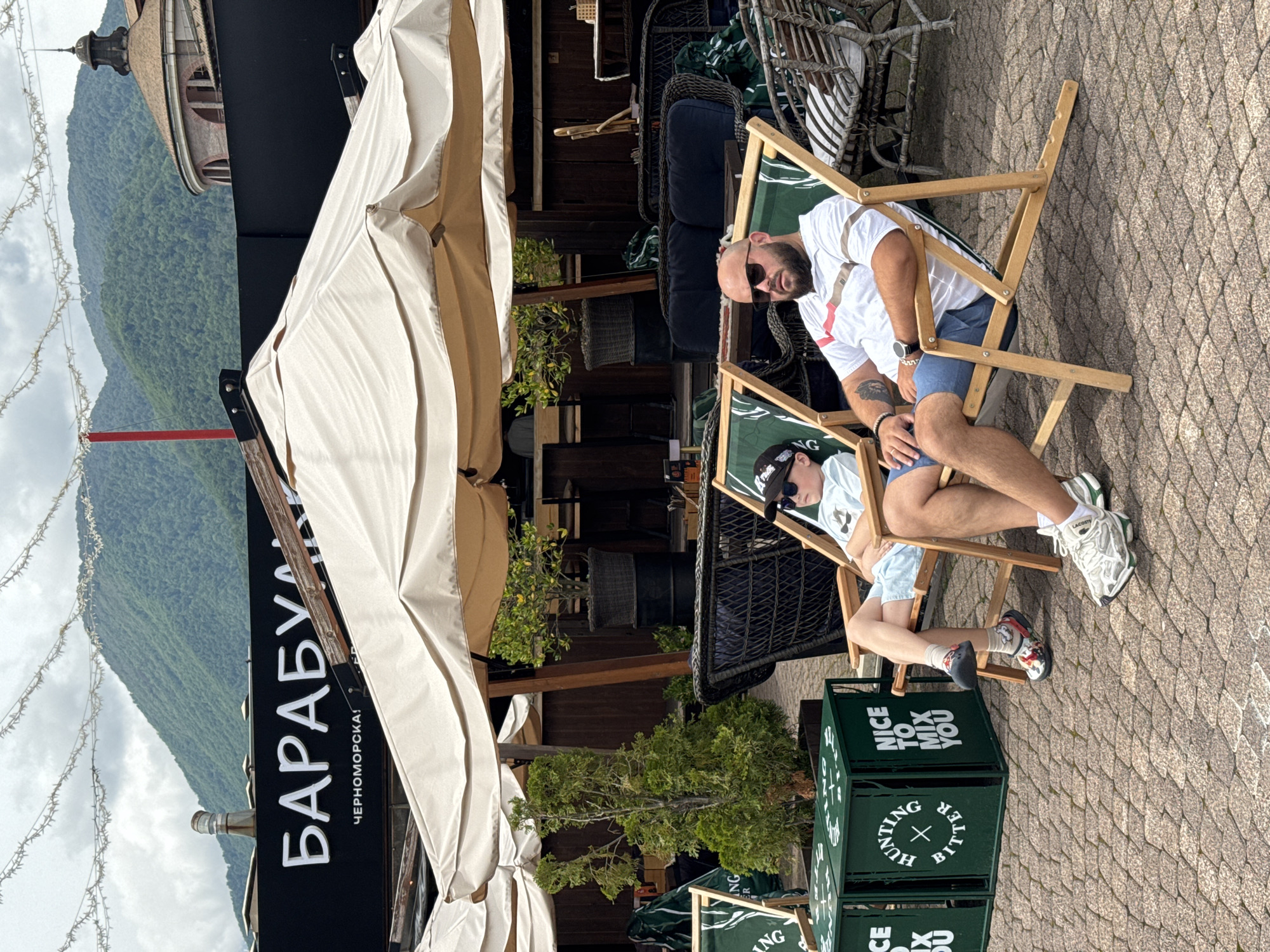 In Estosadok, Russia, a man and a boy relax in lounge chairs under beige umbrellas. The man, wearing a white polo shirt with red and brown stripes and blue shorts, has sunglasses and a watch. The boy, in a light blue t-shirt and shorts, wears a baseball cap and colorful clogs. In the background, a black sign with white Cyrillic text reads "БАРАБУЛКА" (Barabulka), with smaller text below that appears to say "ЧЕРНОМОРСКАЯ" (Chernomorskaya). Green crates with white text, including "HUNTING BITTER" and "NICE TO MIX YOU", are positioned to the left. The scene is set against a backdrop of lush green mountains and a cloudy sky.