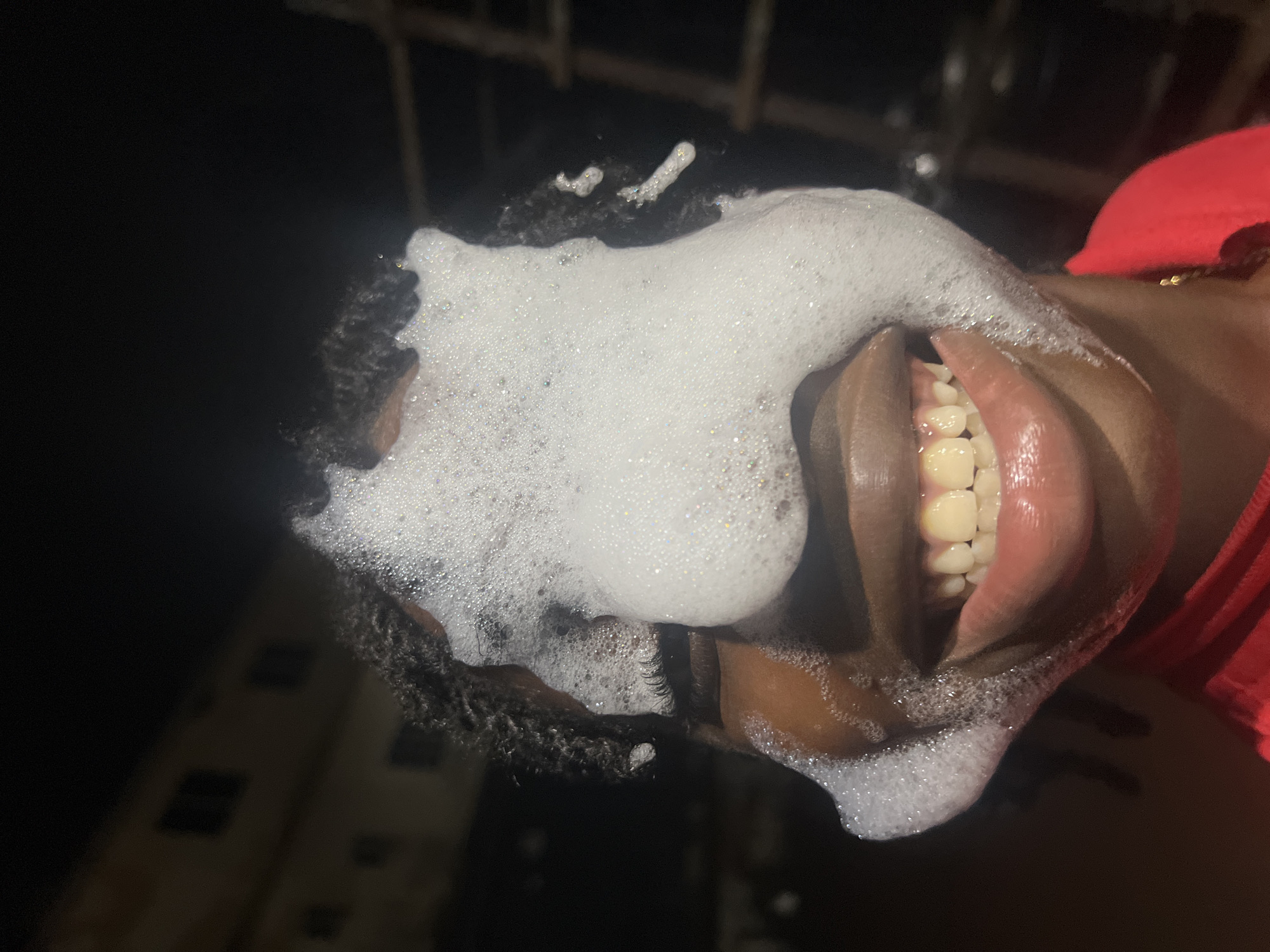 The image shows a close-up, eye-level view of a person's face, partially covered in white foam. The person's eyes are not visible, but their mouth is open in a wide smile, revealing teeth. The foam is spread across the upper part of their face, obscuring their nose and forehead. The person has dark, braided hair. They are wearing a red shirt. The background is dark, suggesting it is nighttime. In the background, a dimly lit building and a ladder or scaffolding structure can be seen. The overall impression is of a playful or experimental moment, likely occurring outdoors at night. The location is specified as Nkpor, Nigeria.