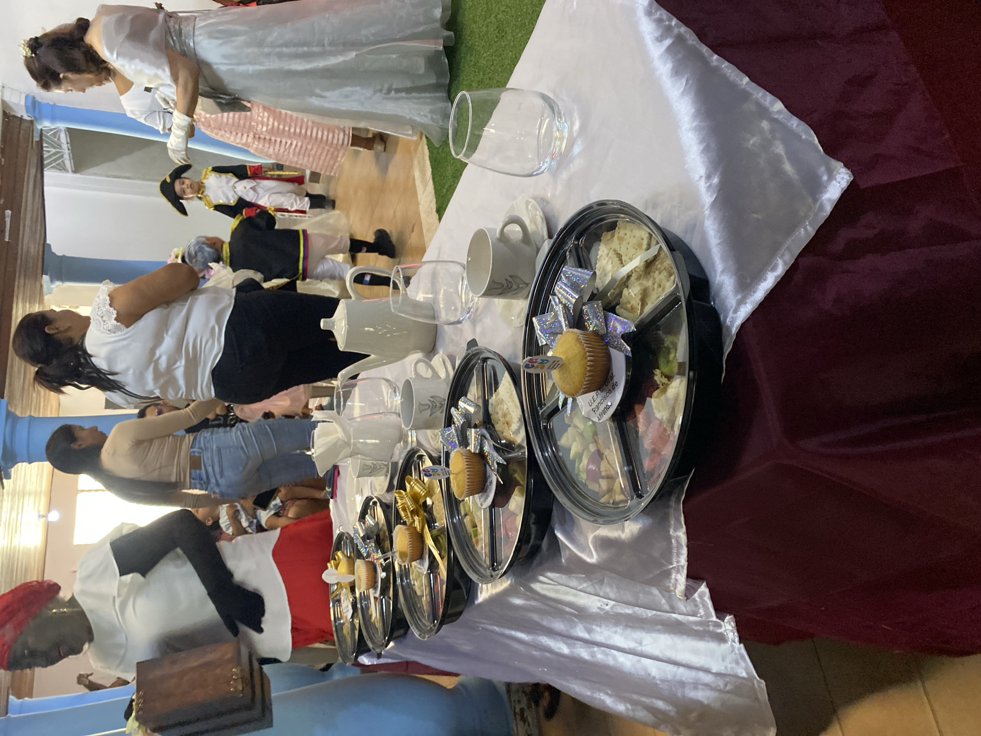 This image depicts an indoor gathering in Tinaquillo, Venezuela. In the foreground, a long table is covered with a burgundy tablecloth and a white satin runner. Arranged on the table are five black, circular, compartmentalized trays, each containing a fruit salad, crackers, and a cupcake adorned with a decorative bow or pick. Also present on the table are white teacups with saucers, clear stemless glasses, a white teapot, and a white pitcher.
Multiple individuals are visible in the midground and background. On the far left, a woman with dark skin wears a red headwrap, a white ruffled blouse, a red skirt, and black gloves, holding a dark wooden box. In the center-left, another woman is partially visible, wearing a light brown top and blue jeans. To the right, a woman in a formal white gown with a silver sash, white gloves, and a hair clip is visible. In the background, two young boys are dressed in historical costumes; one wears a black, white, and red uniform with a bicorne hat, and the other a black jacket with yellow and blue trim, white breeches, and black boots. The room features light blue structural pillars, a tiled floor, and a section of artificial green turf.