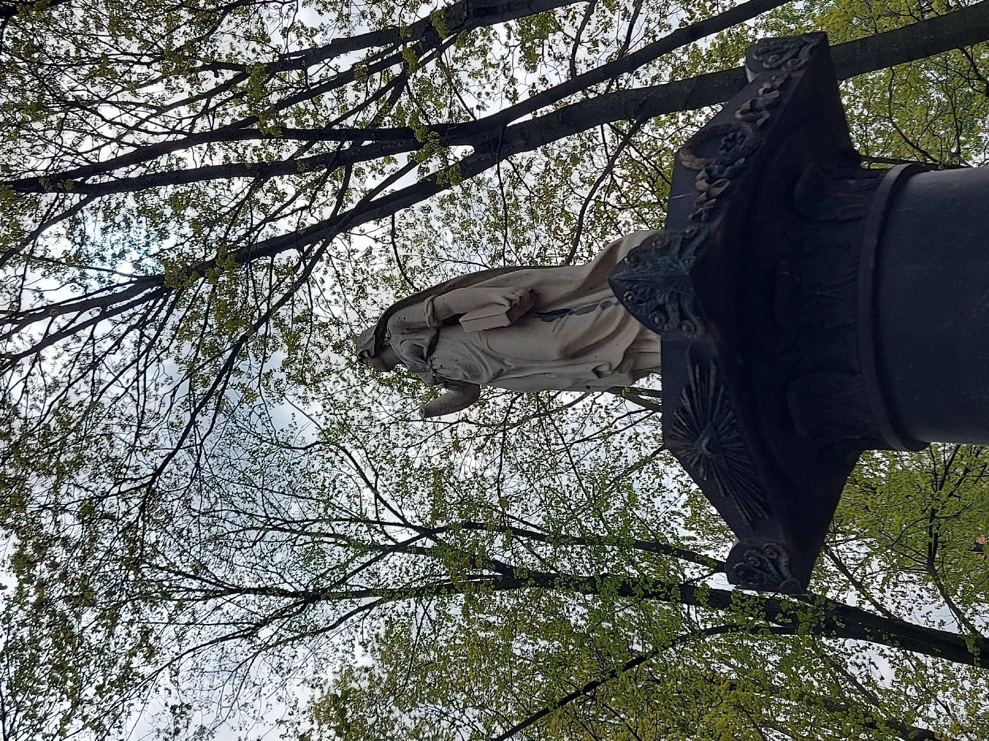 A low-angle shot captures a statue of a figure, likely a religious figure, atop an ornate pedestal, framed by bare tree branches with burgeoning green leaves against a pale sky. The statue is weathered and appears to be made of a light-colored material. The pedestal is dark, possibly metal or stone, with elaborate decorative elements. The location appears to be a park or garden setting in Saint Petersburg, Russia.