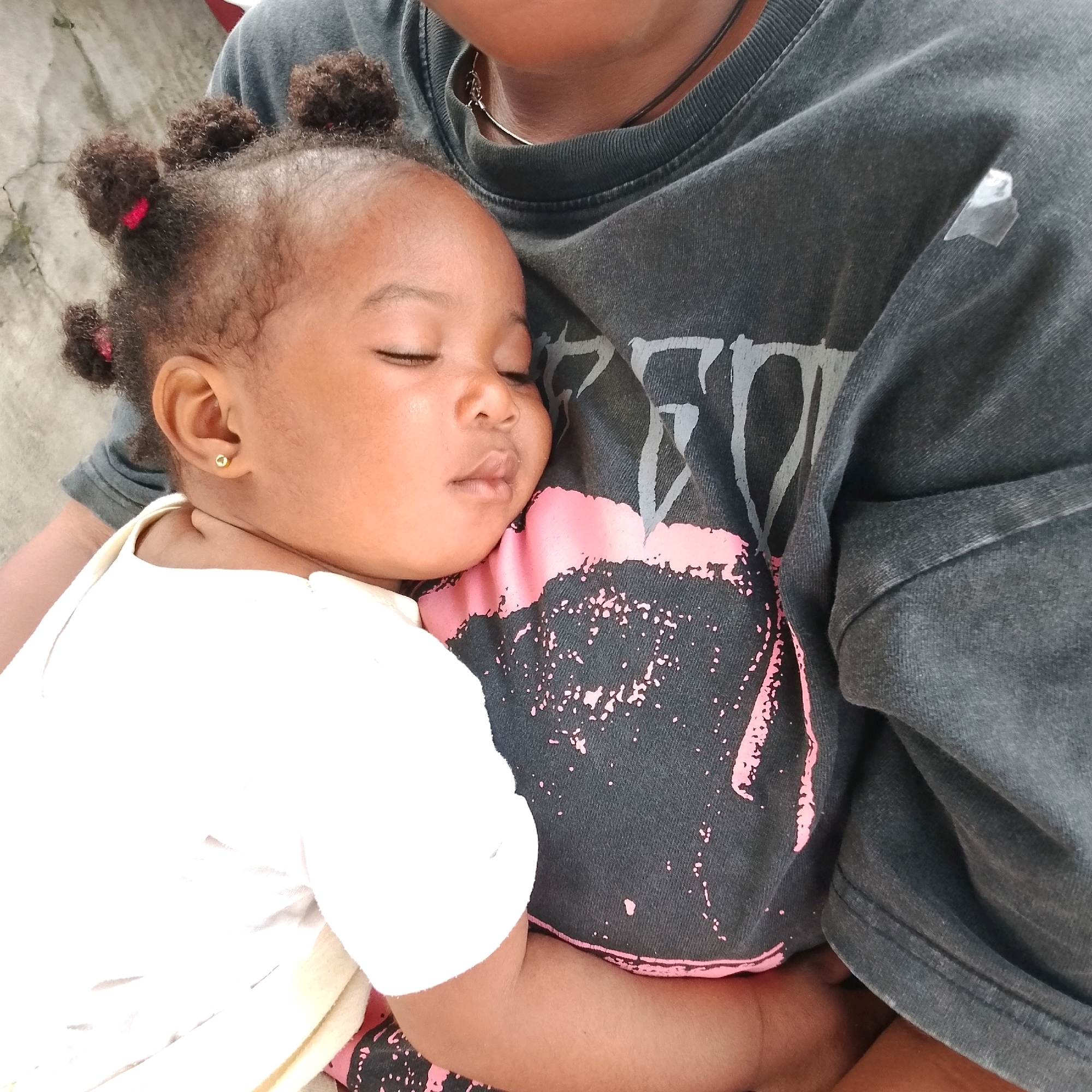 The image shows a close-up of a baby sleeping on an adult's chest. The baby has dark, curly hair styled in several small buns and is wearing a white onesie with a gold stud earring in its left ear. The adult is wearing a dark gray t-shirt with a graphic design featuring pink splatter-like marks. The background appears to be a textured gray surface, possibly a concrete wall. The overall lighting suggests an outdoor or well-lit indoor setting. The scene conveys a sense of intimacy and peacefulness. There are no specific details in the image that can be used to determine its location in Ado, Nigeria.