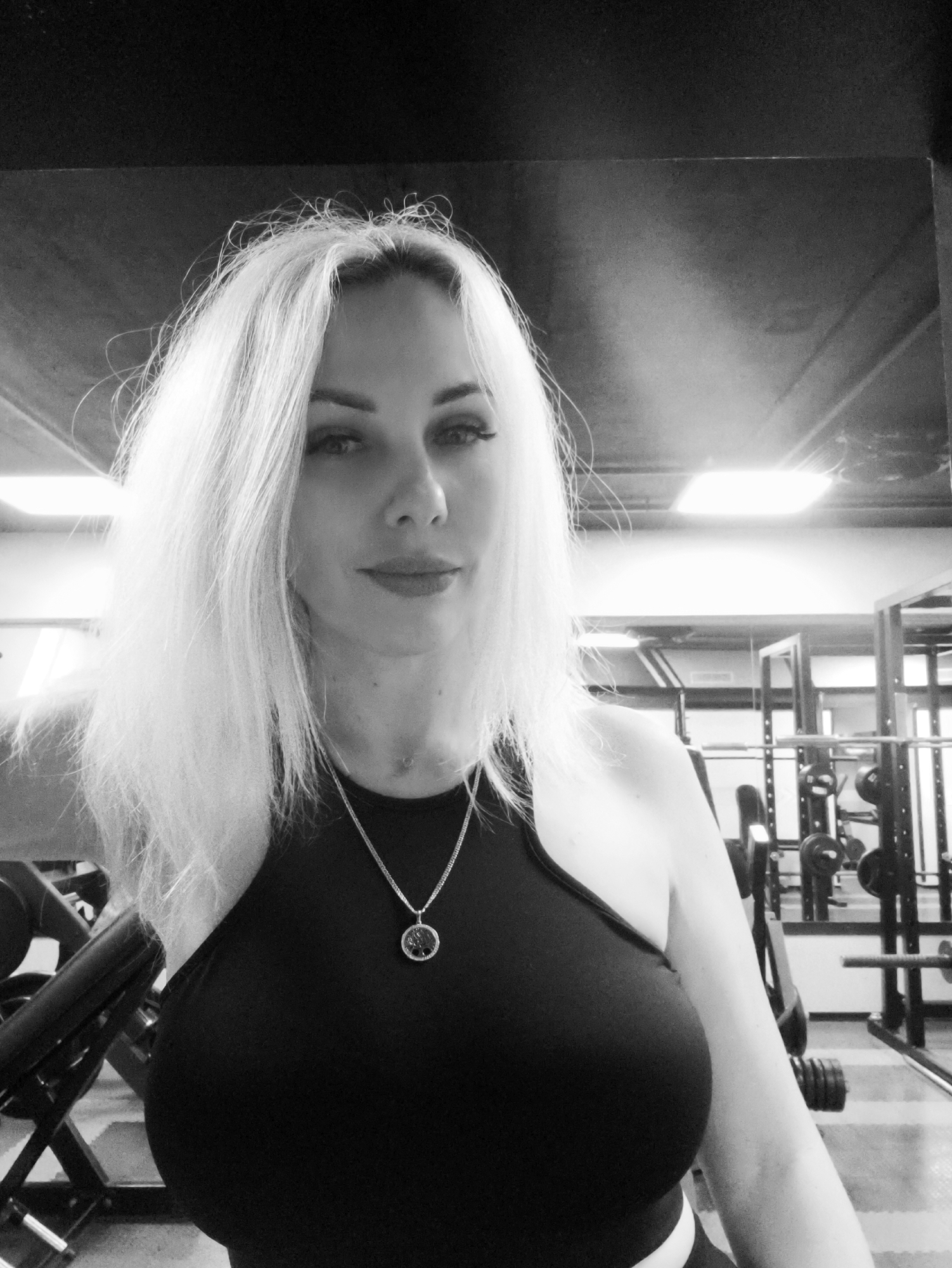 A black and white portrait features an adult woman in an indoor gym setting. She has long, light-colored hair that falls around her shoulders, with noticeable roots. Her eyes are adorned with dark eyelashes, and her full lips show a hint of a smile. She is wearing a dark, possibly black, halter-neck top and a silver-colored chain necklace with a round, detailed pendant. The woman looks directly into the camera with a composed expression.
In the background, various pieces of gym equipment are visible, including squat racks, weight plates, and what appear to be dumbbells and benches. Reflections of the equipment and rectangular overhead lights can be seen in mirrors along the back wall, clearly indicating a fitness facility. The image is a static shot, likely a posed selfie, with no active event taking place beyond the woman presenting herself in the gym environment. There is no visible text.