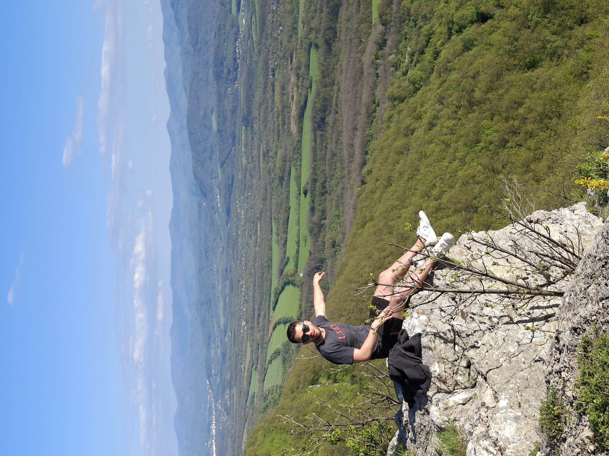 A man stands on a rocky cliff, likely in the Zdravkovets region of Bulgaria, overlooking a valley with forests and fields. He wears a grey shirt, black shorts, sunglasses, and white sneakers. His left arm is raised. A black jacket or bag sits beside him on the rocks. The background features a blue sky and a mountainous landscape.