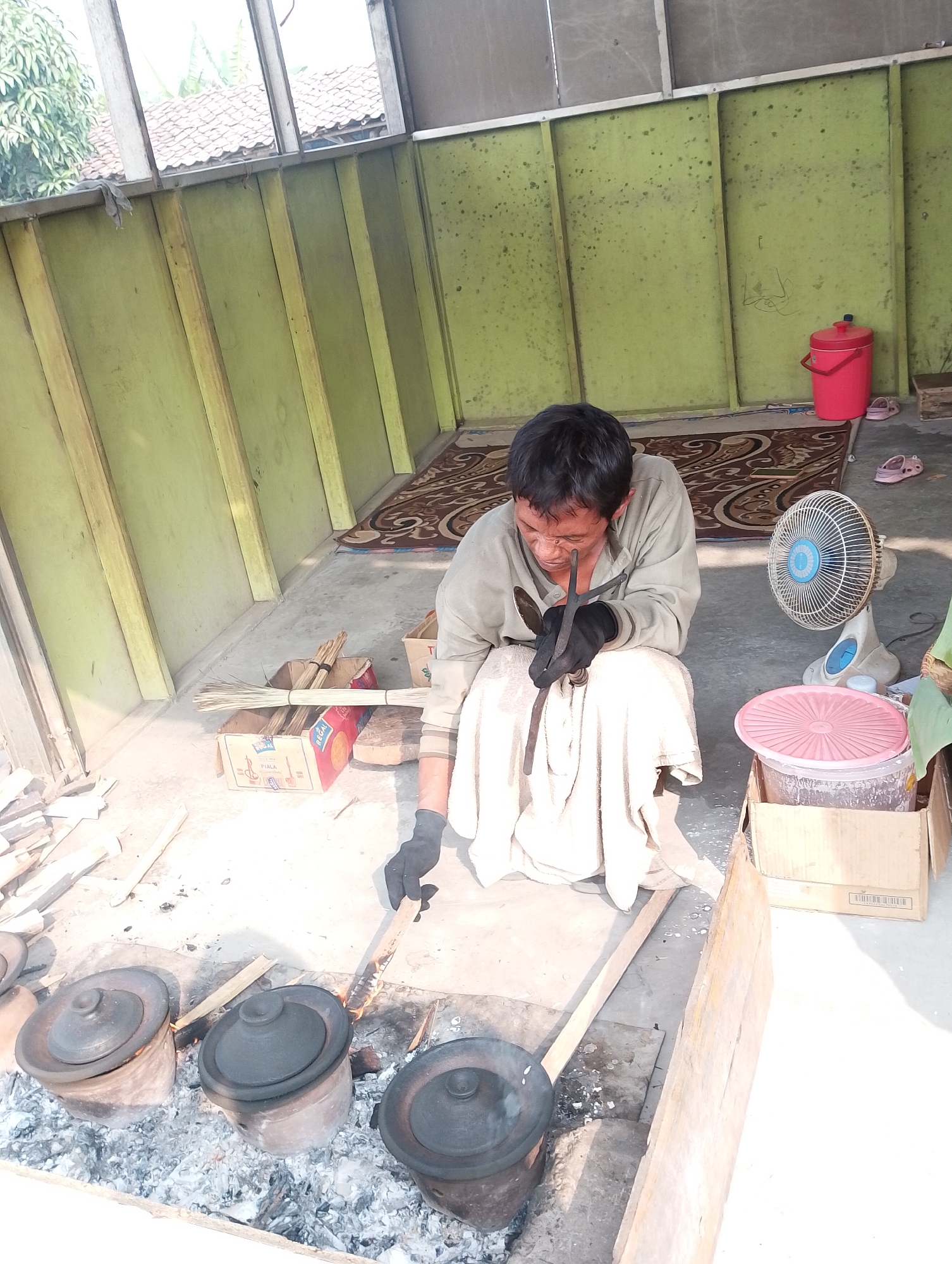 The image depicts an outdoor workshop or cooking area in Karawang, Indonesia. A man wearing a light-colored shirt and black gloves is seated on the ground, tending to a fire where several clay pots are placed. He is holding a tool and appears to be engaged in some form of crafting or cooking. A fan is positioned behind him, and various items like brooms, boxes, and a red bucket are visible in the background. The environment suggests a rustic and possibly informal setting.