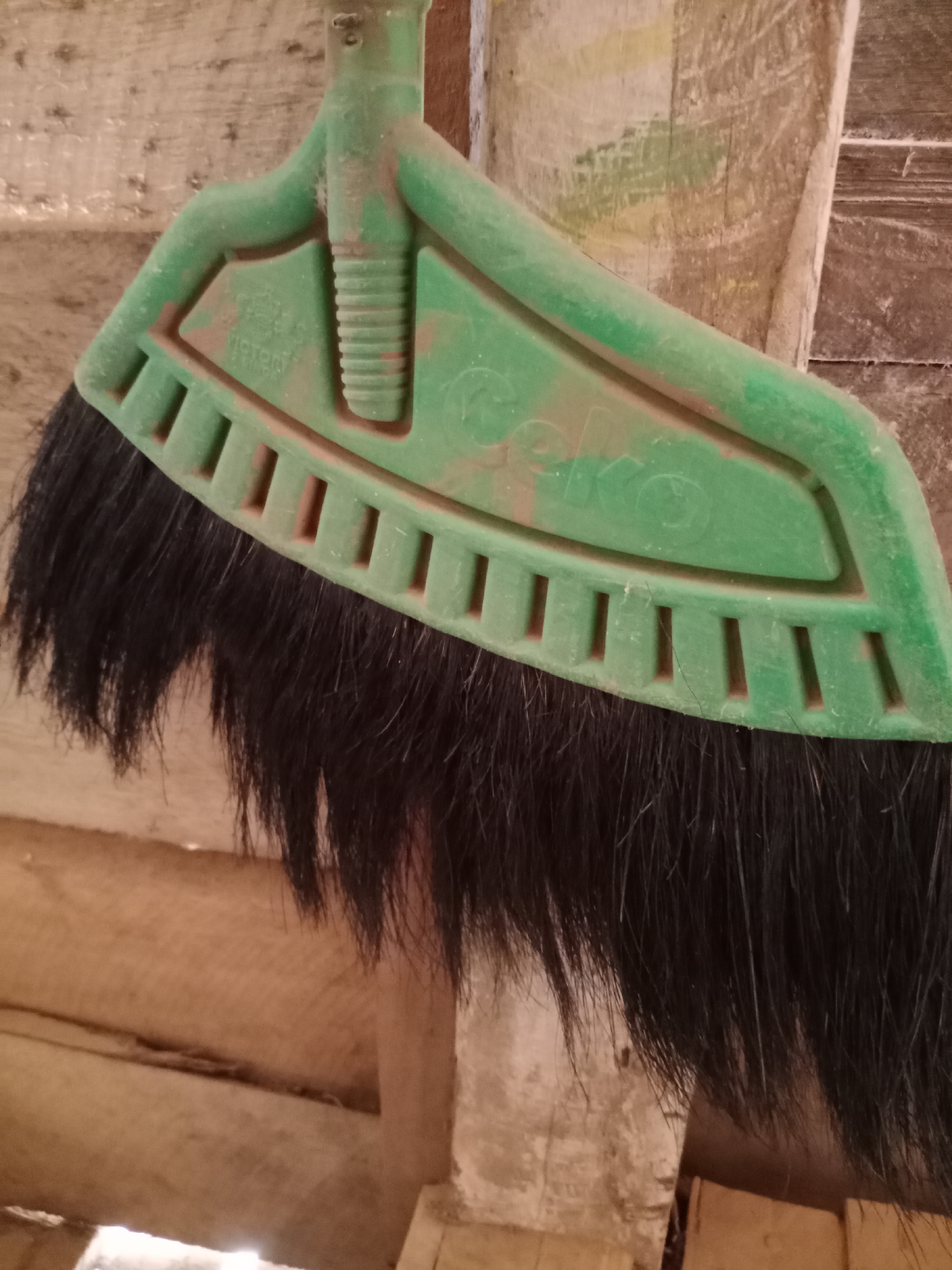 A green plastic broom head with black bristles is hanging on a wooden structure. The broom head has some raised text, but it is mostly obscured by dust. The wooden structure appears to be made of rough-hewn planks, some of which are weathered and stained. The overall scene suggests an outdoor or rustic indoor environment.