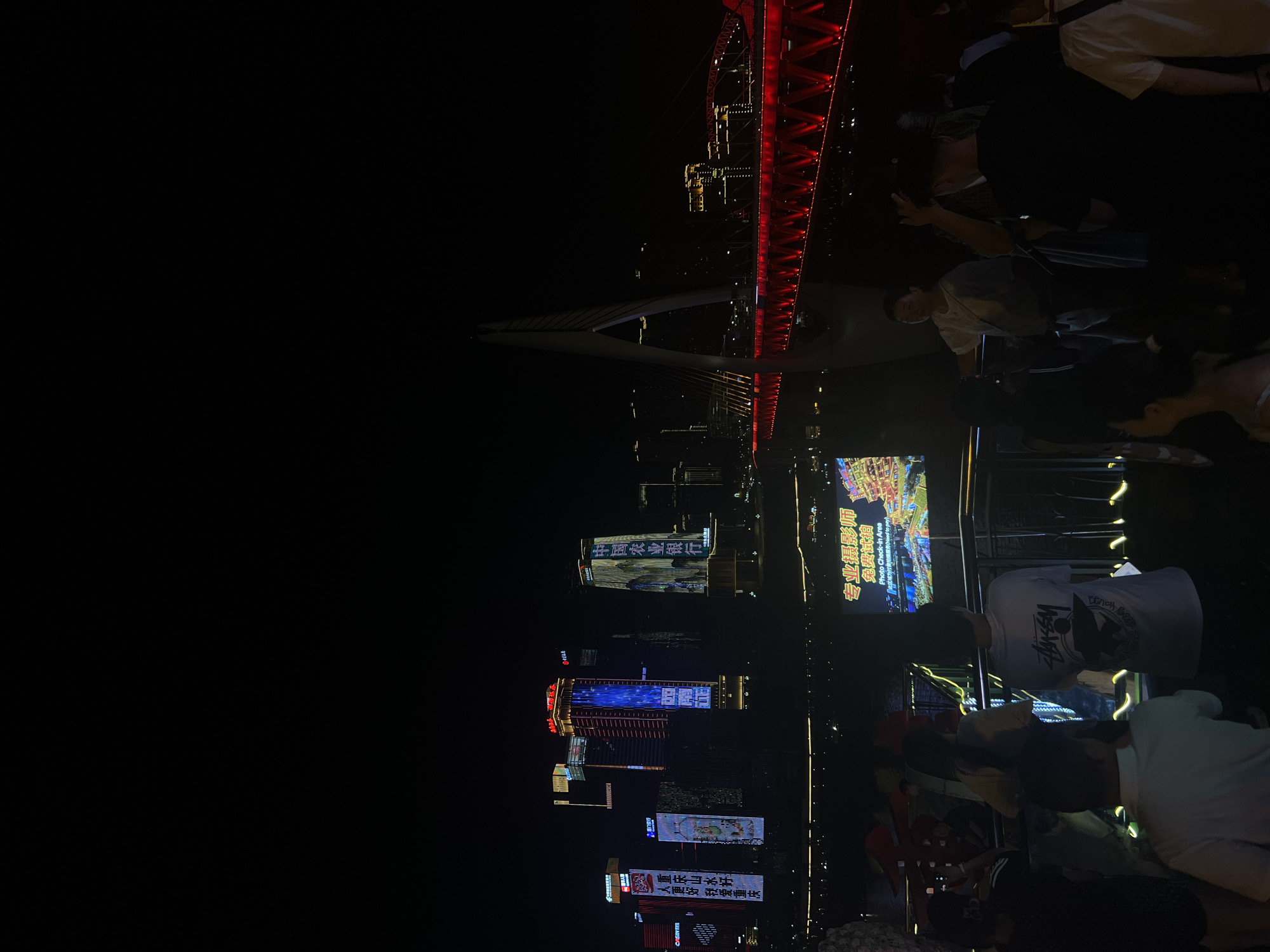 This is a nighttime outdoor photograph taken in Yuzhong District, China, likely on a waterfront promenade overlooking a river and illuminated city skyline. In the foreground, several people are standing and looking out towards the view. One person on the right is wearing a white t-shirt with a graphic print. To the left and center, other individuals are visible, some in casual clothing.
The midground features a railing or barrier, suggesting a viewing area. Beyond this, a large screen displays an animated city map with Chinese text. In the background, a modern suspension bridge with red lighting spans the river, with a tall, white pylon. The far background shows a cluster of illuminated skyscrapers, some displaying vertical LED advertisements. The overall impression is a vibrant urban night scene with people enjoying the cityscape.