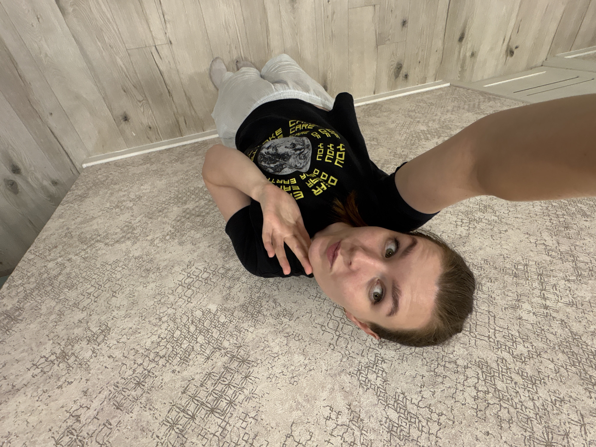 A woman in a black t-shirt and grey sweatpants lies on a carpet with a patterned design. Her head is towards the lower right corner of the image, and her legs are in the upper left corner. The walls are light wood paneling. The photo appears to be a selfie taken by the woman, who is looking at the camera. The location appears to be an interior space. Izhevsk, Russia is not discernible.