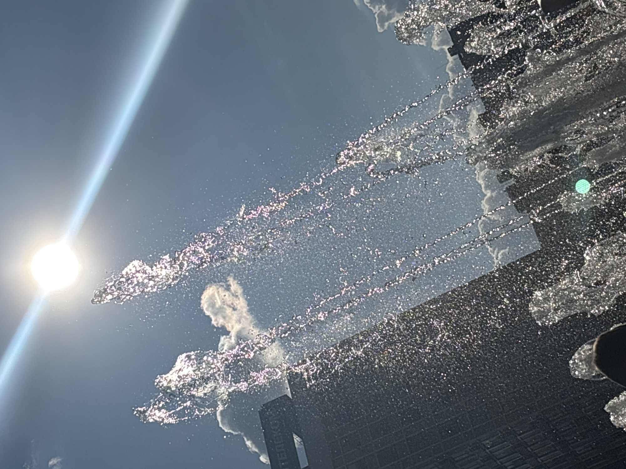 The image shows a fountain spraying water in the foreground against a backdrop of a bright sun and a blue sky with clouds. The sun is positioned in the upper left quadrant of the image, with a lens flare extending diagonally across the sky. Behind the fountain, a building with a sign that reads "ЕЛЕКС" is visible on the left side, and other buildings form the city skyline. The water from the fountain is illuminated by the sun, creating a sparkling effect. In the bottom left corner, a person's head and shoulders are partially visible, suggesting they are standing near the fountain. The overall scene depicts an urban environment on a sunny day.