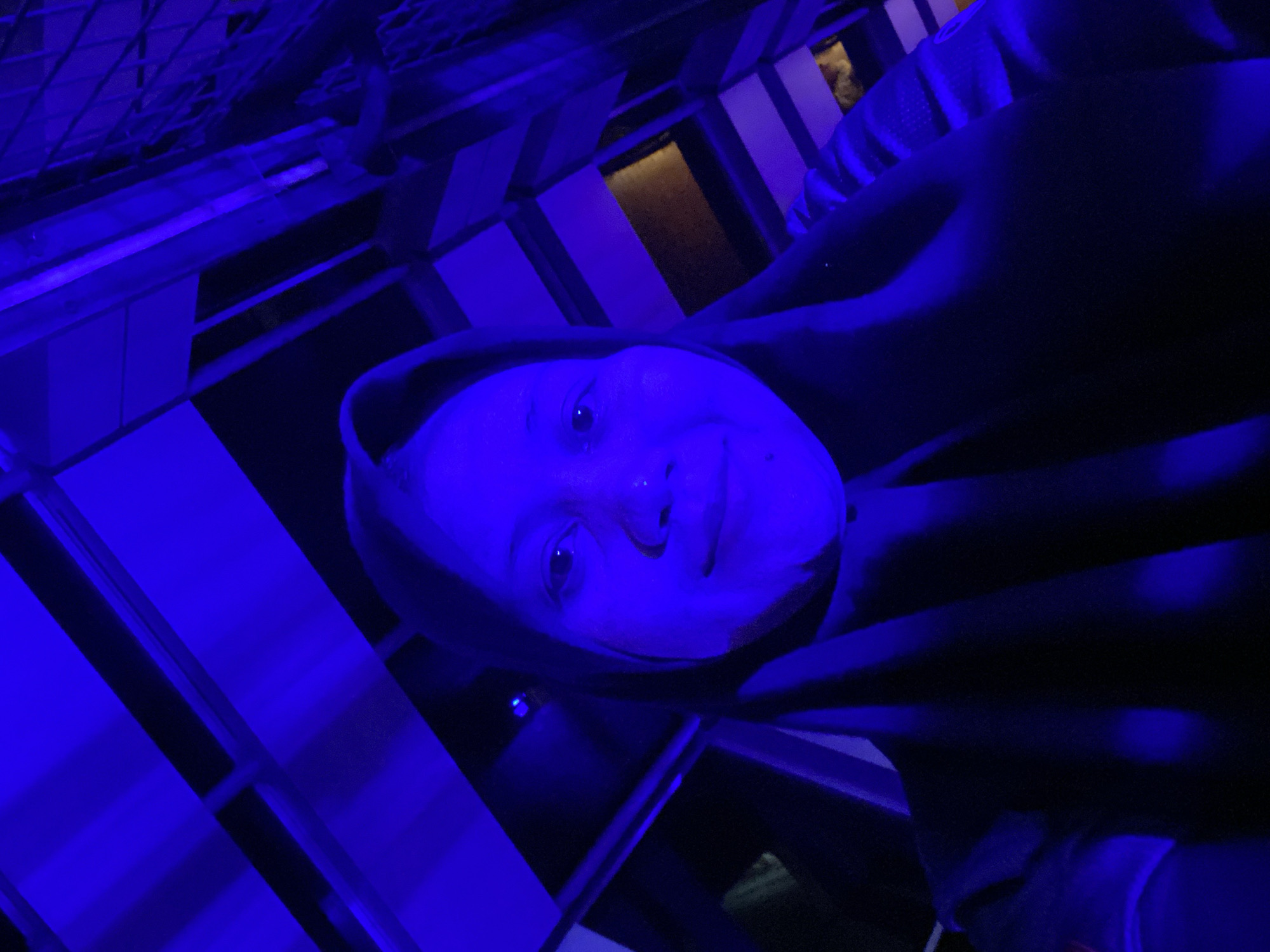 A selfie taken by a young woman wearing a dark hijab and dark clothing, illuminated by bright blue lighting. She is looking directly at the camera with a neutral expression. The background is blurred, showing a structure of diagonal and horizontal beams, likely part of a building's exterior or interior. The overall impression is that the photo was taken at night or in a dimly lit area with artificial blue lighting. The location is identified as Banjarbaru, Indonesia.