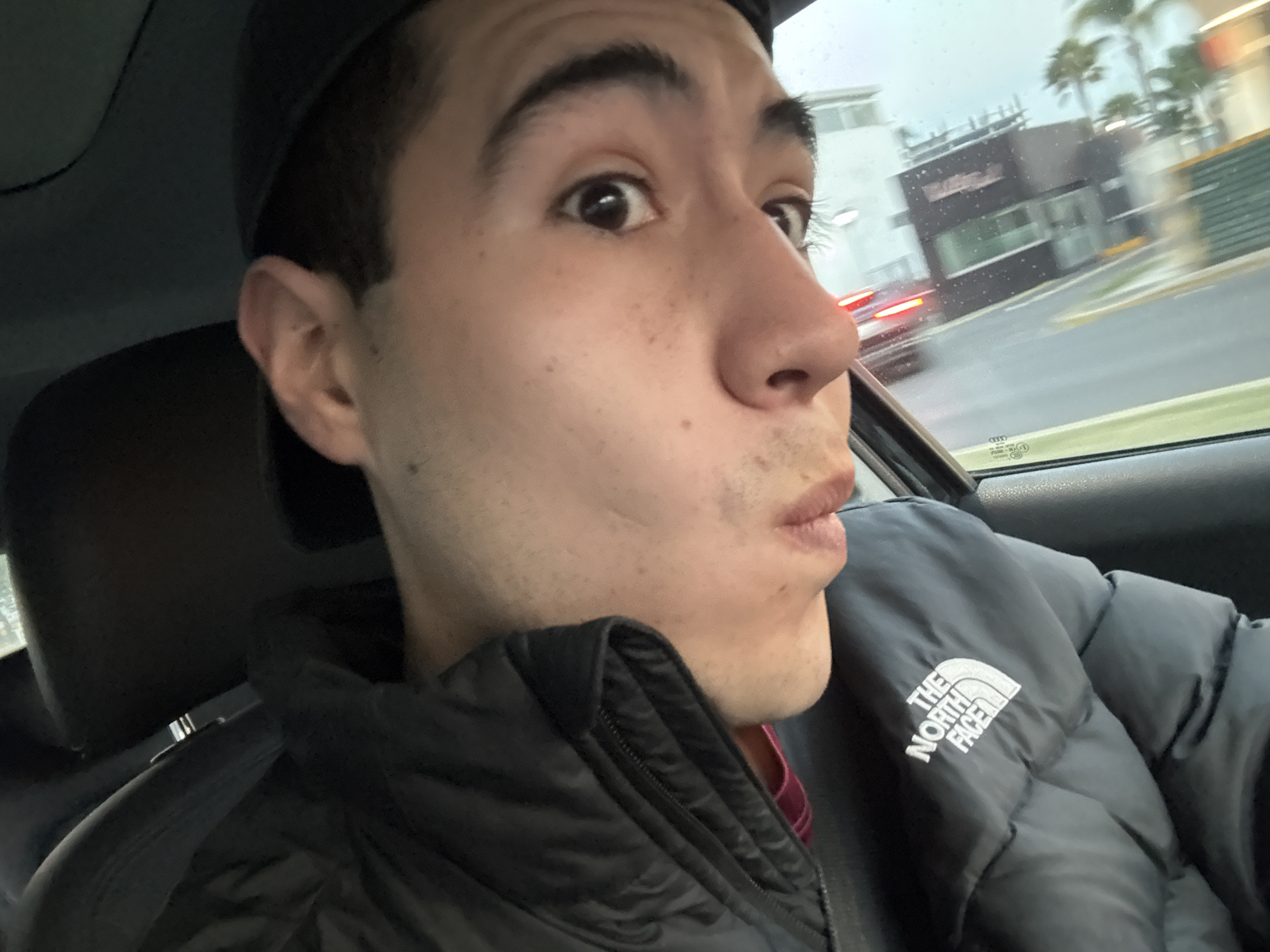 A light-skinned male with dark hair is in the driver's seat of a vehicle. He is wearing a black baseball cap and a black puffy jacket with the North Face logo on the left side. His face is turned toward the passenger side with a surprised expression. Outside the window, a building and palm trees are visible along with cars moving in a blurred motion, suggesting the vehicle is in motion. The rain is visible on the front window. The scene takes place in the city of San Bernardino Tlaxcalancingo, Mexico.