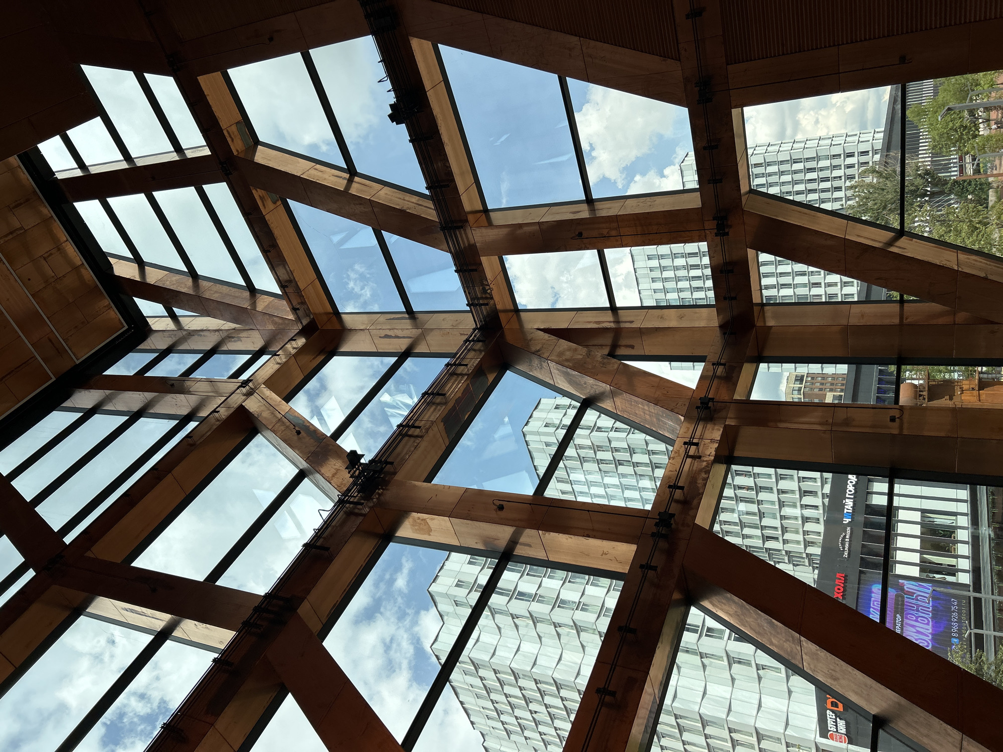 The image captures an interior view of a modern building, characterized by its intricate wooden structural beams and large glass panels. The wooden beams form a complex geometric pattern that extends upwards and across the frame, creating a sense of depth and architectural complexity. Through the glass, a cityscape is visible, featuring several tall, modern buildings with a predominantly white or light-colored facade and numerous windows. The sky above is a bright blue with scattered white clouds, indicating it is daytime. Some of the visible building facades appear to have a terraced or stepped design. Below the main buildings, there are also glimpses of lower structures and greenery, suggesting a partially urbanized environment. Signs are visible on some of the lower buildings, including what appears to be a Burger King sign. The overall impression is one of contemporary architecture, blending natural materials with modern design, set against an urban backdrop. The perspective is from within the structure, looking outwards through the expansive windows. There are no people visible in the image.