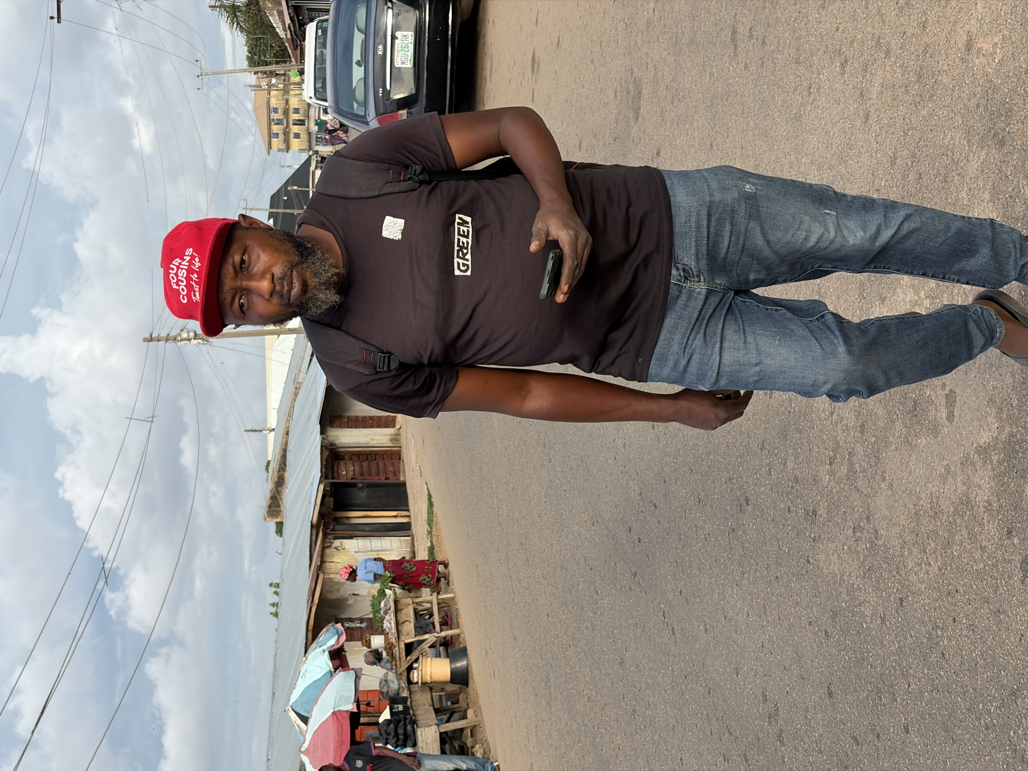 The image depicts an outdoor street scene in Jos, Plateau State, Nigeria, under an overcast sky. A dark-skinned male individual, with a beard and mustache, walks in the foreground towards the viewer's right. He wears a red baseball cap with "FOUR COUSINS Taste to Life!" in white text, a black t-shirt with a white "GREEN" logo and a square emblem above it, blue jeans, and dark flip-flops. A black backpack is on his shoulders. He holds a dark-colored smartphone in his left hand and looks directly towards the camera.
To the left, market stalls with merchandise and two people are visible. One person is partially obscured under a red and light blue striped umbrella. Another female figure, wearing a red patterned skirt, light blue top, and pink head covering, stands near a wooden stall displaying green produce. Behind the stalls are single-story buildings with corrugated metal roofs.
On the paved road to the right, a black Kia sedan is parked, displaying the Nigerian license plate "MGU-262AK", which is consistent with Plateau State registration. In the background, a multi-story yellow building and utility poles with overhead wires are present.