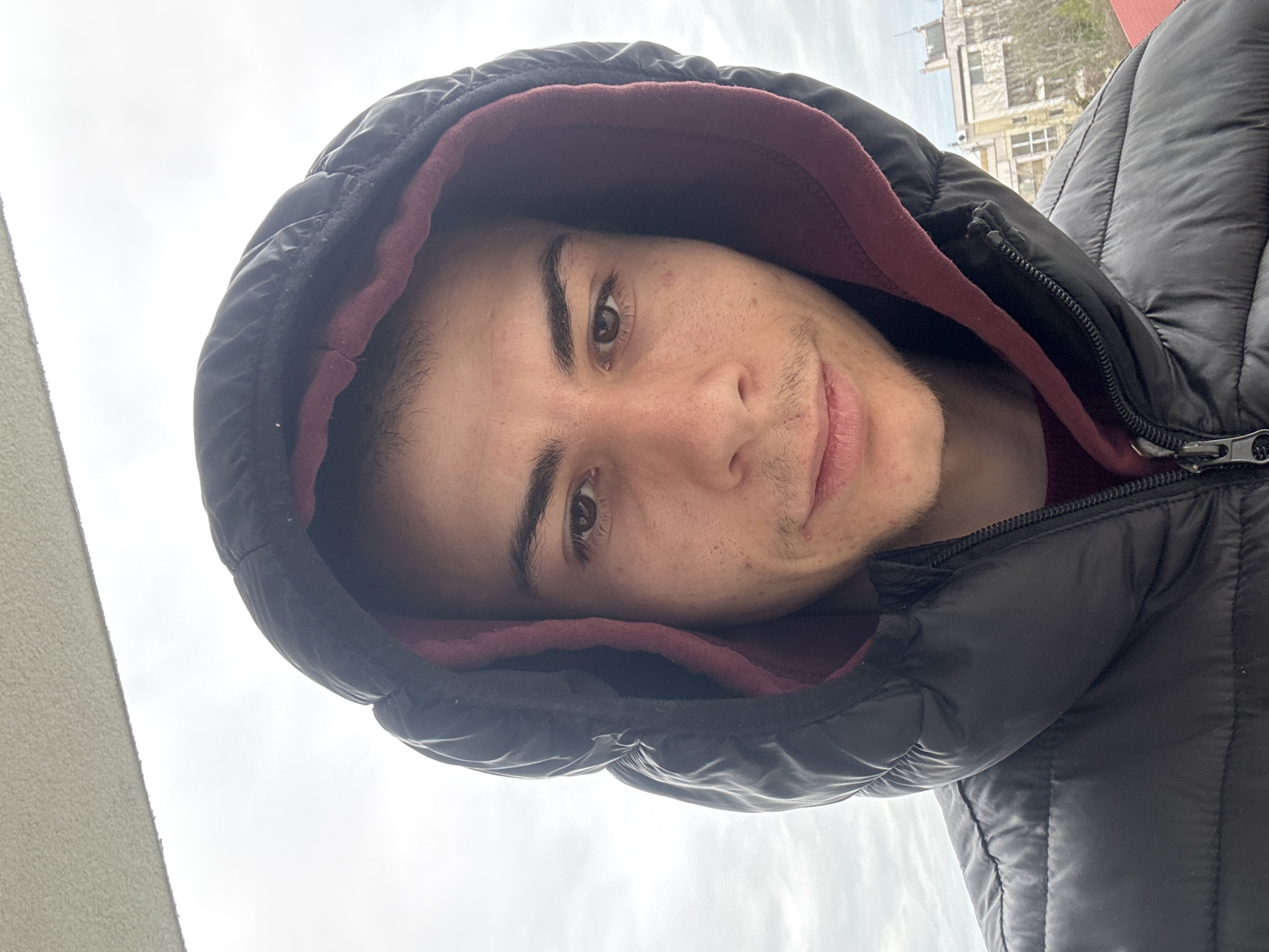 A young man with fair skin and brown eyes is centered in the image, wearing a black puffy hooded jacket with a maroon lining. He has dark eyebrows and slight stubble on his face. The hood of the jacket is up, framing his face. The background features a cloudy sky and a building with multiple windows, potentially located in an urban setting. Based on architectural style, this may be located in the city of Plovdiv, Bulgaria.