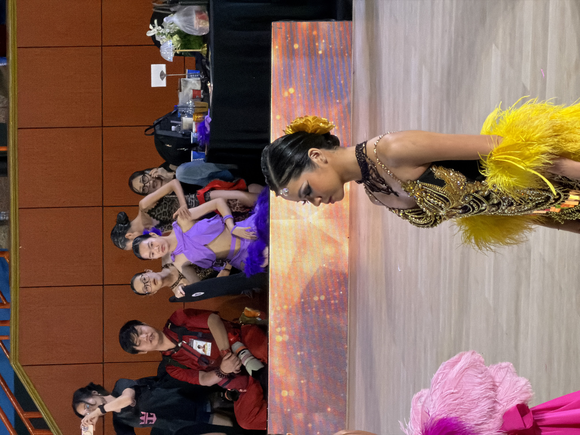 The image captures a dance competition or performance with participants in elaborate costumes on a stage. In the foreground, a young woman with dark hair styled in an updo, adorned with a yellow flower, is shown in profile, wearing a black and gold sequined dress with yellow feather accents. Behind her, another dancer in a bright pink and fuchsia feathered costume is partially visible on the left. In the middle ground, a group of people are seated, including a man in a red shirt holding a camera and a young woman in a purple and blue costume. The background shows a stage with a vibrant, illuminated screen. The setting appears to be an indoor venue, likely a theater or auditorium, with tiered seating visible at the top. The overall atmosphere suggests a lively event. The specific location of this scene in Hà Nội, Vietnam, cannot be determined from the image alone.