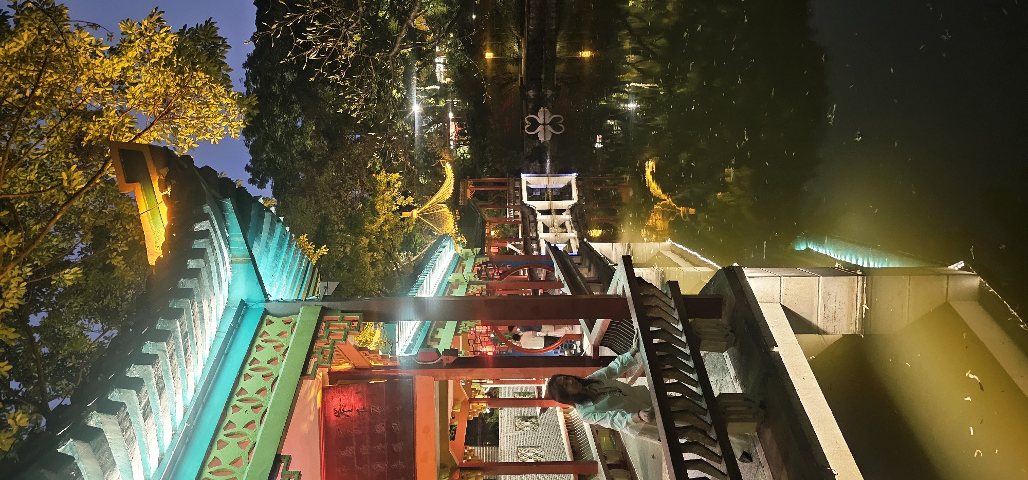 A nighttime scene in Yuzhong District, China, shows a person in light-colored clothing standing on a walkway adjacent to a body of water. The walkway is decorated with red and brown wooden beams and a green roof illuminated with blue and yellow lights. The water reflects lights from nearby buildings and trees. A structure with a white frame and a cloverleaf design sits further away over the water. In the background, trees frame the scene, some illuminated with strings of lights.