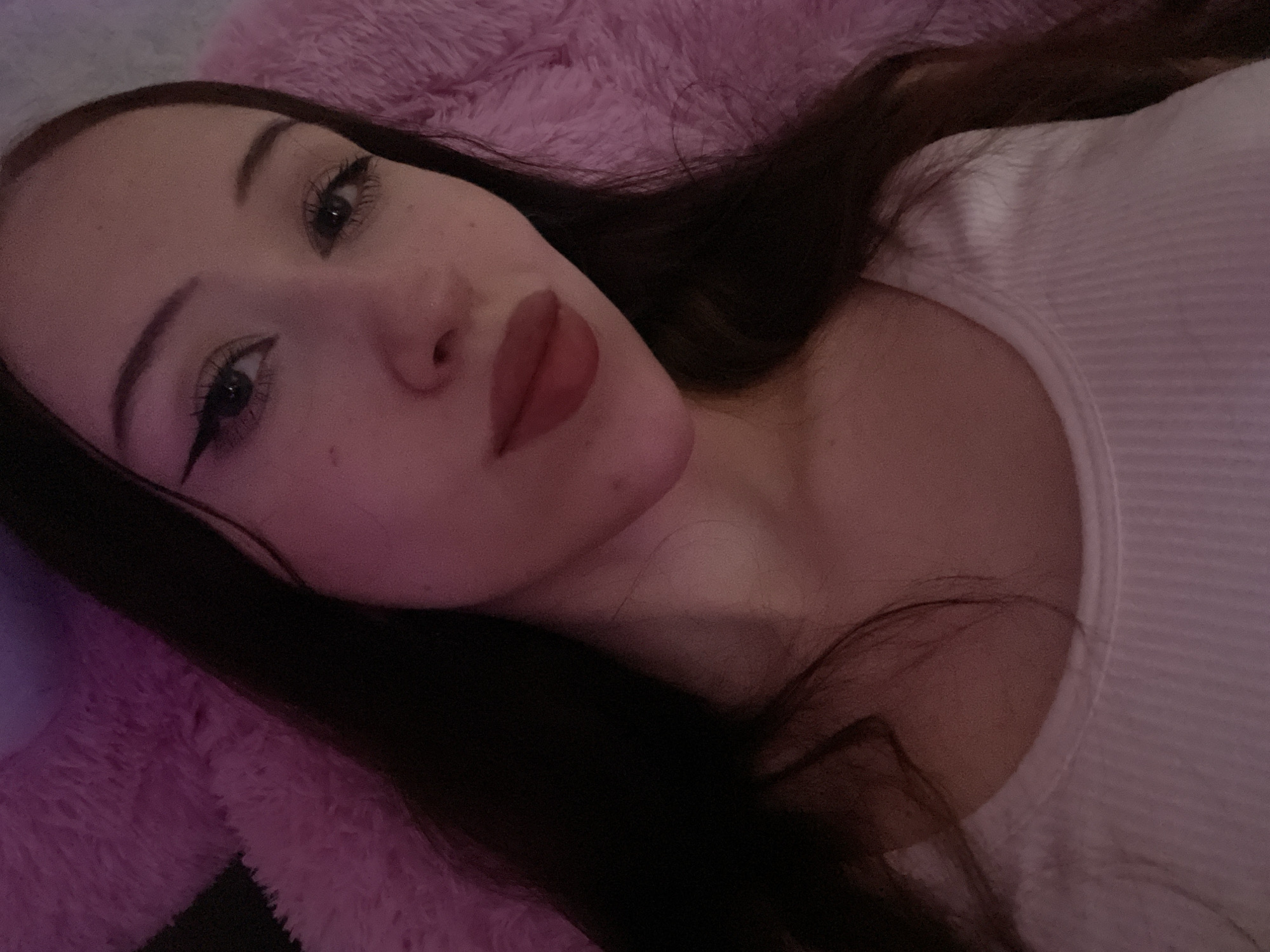 The image shows a close-up, selfie-style shot of a young woman with long, dark brown hair. She is wearing a white ribbed tank top. Her makeup includes winged eyeliner and lipstick. She is positioned against a blurred background of a pink, fluffy surface, possibly a blanket or stuffed animal. The lighting is dim and appears to have a pinkish hue. There is no identifiable location within the city of Votkinsk, Russia.