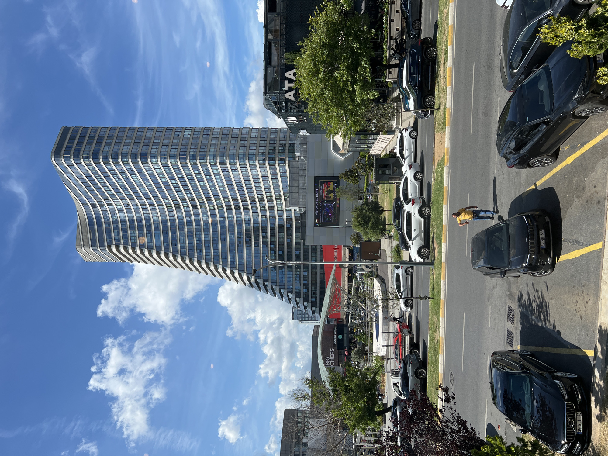The image captures a street scene in Ataşehir, Turkey, featuring a distinctive modern skyscraper with a curved, glass facade that tapers towards the top. Adjacent to the skyscraper is a dark-colored building with the prominent white lettering "ATA" on its facade, suggesting a business or commercial establishment. In the foreground, several cars are parked on the street and in a lot, including black sedans and SUVs. A man in a yellow shirt and blue jeans is walking across the street. Further down the road, white and silver cars are moving, indicating active traffic. The background reveals a bright blue sky with scattered white clouds and greenery from trees. A large digital screen is visible on one of the buildings, displaying information. The overall impression is of a dynamic urban environment on a clear, sunny day.