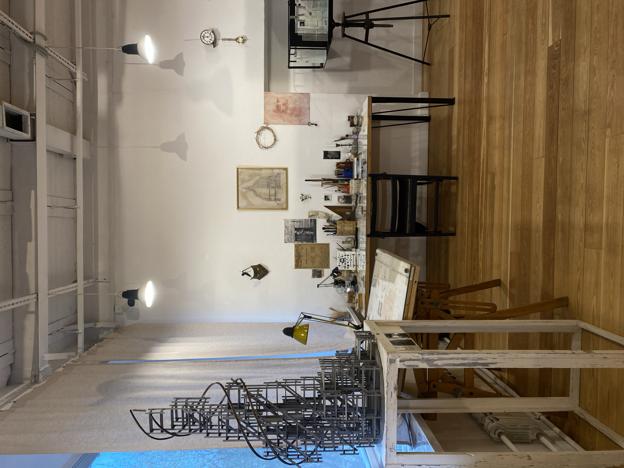 This media file captures an indoor setting, likely an artist's or architect's studio, workshop, or a curated exhibition designed to resemble one. No people are visible.
The scene features a light-colored wooden floor and white walls. The ceiling has exposed white structural beams with modern spotlights casting prominent shadows. Natural light enters from a window on the left, but artificial lights are also on.
Central to the image is a large wooden drafting table with a tilted surface, laden with papers and tools. To its left, a complex metal architectural model or sculpture, composed of geometric and curved elements, stands prominently. In the foreground, a distressed white wooden frame structure adds to the artistic ambiance. Further into the room, against the far wall, is a long wooden table with black legs, densely covered with various art and drafting supplies including brushes, pencils, rulers, small sculptures, and magnifying glasses. A wooden chair is tucked beneath this table. On the far right, a wooden easel supports a display case containing what appears to be another architectural model.
The white walls are adorned with numerous items. Two clocks are visible, one a classic round wall clock, the other a smaller, more ornate clock. Various framed and unframed sketches and papers are hung, including a detailed drawing of a classical dome in a gold frame and a reddish-toned sketch of a figure. A "crown of thorns"-like object and a single key are also mounted on the wall. Several pieces of aged paper with faint, mostly illegible text and handwritten notes contribute to a collage effect. One piece might read "TRADIZIONI."
The setting conveys a sense of creative activity and dedication to craft or study, possibly historical, given the style of some artifacts. The location context is Moscow, Russia, although no specific cues within the image confirm this.