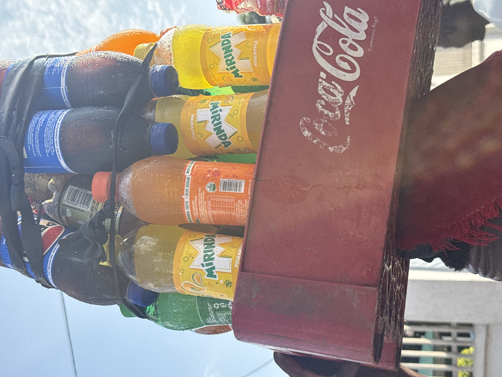 This is a close-up, low-angle, outdoor shot of a stacked arrangement of beverages, with a focus on a red Coca-Cola crate. The crate is positioned at the bottom of the frame and bears the white Coca-Cola logo, with "Trademark Regd" printed below it. Above the crate, several plastic bottles of soft drinks are stacked vertically.
The drinks visible include:
- Two bottles of Mirinda Pineapple, identifiable by their yellow labels and the word "MIRINDA" in white lettering.
- One bottle of what appears to be Fanta Orange, with an orange-colored liquid and an orange cap.
- One green bottle, likely Sprite or another citrus-flavored soda.
- Two dark-colored bottles, possibly Coca-Cola or Pepsi, with blue caps and labels that have prominent blue sections. These bottles appear to be sweating, indicating they are cold.
The bottles are secured together with black strapping. The background suggests an outdoor environment with a bright sky, possibly with some clouds. Hints of buildings and possibly a fence are visible in the far background, to the left and right.
While the image does not explicitly show the city of Owerri, Nigeria, this type of stacked beverage display in a Coca-Cola crate is a common sight in informal markets and street vending scenarios across Nigeria, including Owerri. The presence of these specific brands and the style of presentation are consistent with such locations in Nigerian cities. There is a hint of a person's dark profile and a red cloth to the right and left edges of the frame, suggesting this display might be part of a larger street scene or market stall.