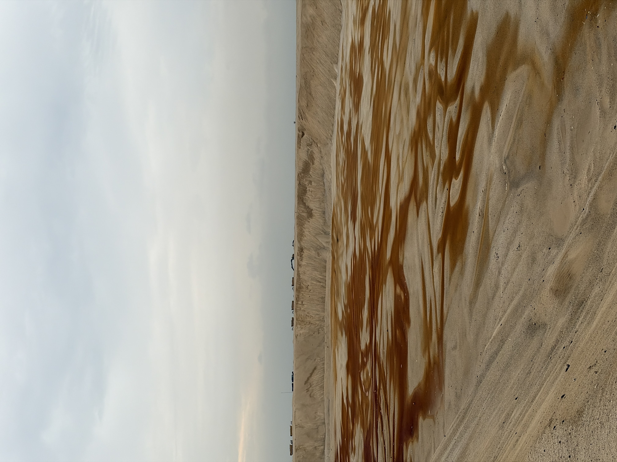 An aerial view of a sand quarry in Alaguntan, Nigeria, shows a vast, excavated pit filled with water and sand. The water, a dark reddish-brown, pools in irregular shapes across the sandy floor, creating a rippled, abstract pattern. The edges of the pit are steep, composed of layered sand formations. Along the rim of the quarry, a line of trucks and construction equipment is visible, suggesting ongoing activity. The sky above is overcast with muted, gray clouds, casting a diffused light over the entire scene. The overall impression is one of industrial extraction and a desolate, arid environment.