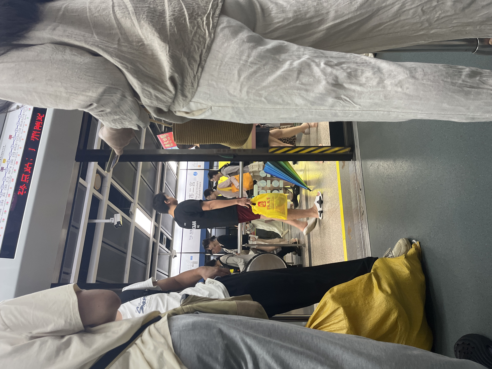An indoor shot captures a group of people at a subway station platform in an unknown city. In the foreground, a person wearing a beige top and light-colored pants stands facing away from the camera. To the left, another individual in a white t-shirt and grey sweatpants is partially visible, with a large yellow bag at their feet.
Further into the scene, a man in a black t-shirt and red shorts walks by, holding a yellow shopping bag. Behind him, several other individuals are seen near a subway entrance, some walking, others standing. A train is visible at the platform edge, and station signage with Chinese characters is displayed overhead. The environment appears to be clean and well-lit, typical of a modern subway station. An open rainbow-colored umbrella is also visible on the platform.