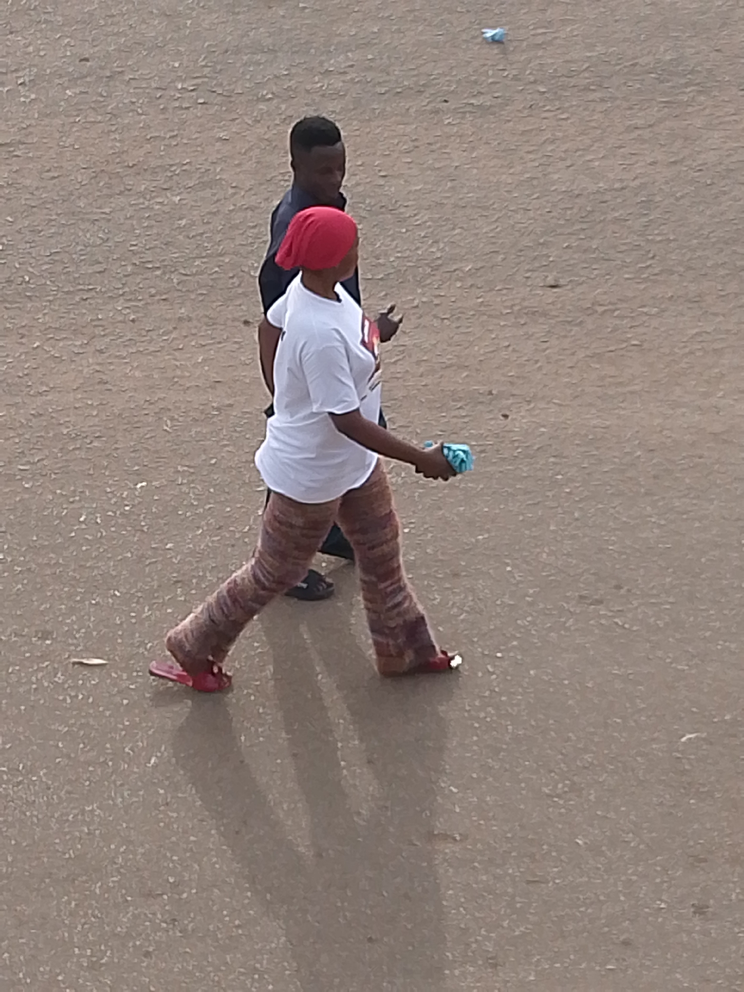 Two individuals are walking on a paved surface, likely a road or pathway. The person in the foreground is a woman wearing a red headscarf, a white t-shirt, and patterned trousers. She is carrying a small blue object in her right hand and wearing red sandals. Behind her and slightly to the left, a man is visible, wearing a dark shirt. The scene appears to be outdoors, and the lighting suggests it could be daytime, possibly overcast. The ground is textured and appears to be asphalt or a similar material, with some small debris scattered on it. There are no visible signs of text in the image. The overall impression is a candid shot of people going about their day in an urban or semi-urban environment in Bukuru, Nigeria.