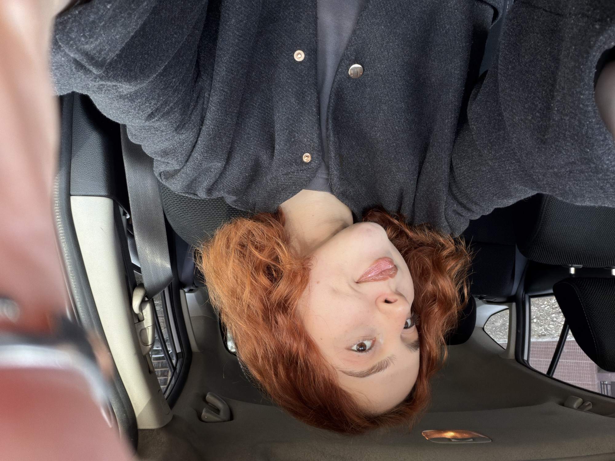 A person with red, curly hair and fair skin is in the passenger seat of a car, possibly in Tbilisi, Georgia (location speculation based on details provided in the prompt). They are wearing a dark gray blazer with gold-colored buttons. The seatbelt is fastened. The view is tilted, showing the car's interior, including the dashboard, seats, windows, and interior lights. The background visible through the windows suggests an urban environment. The person is looking towards the camera, with a slightly playful expression, sticking their tongue out slightly. The lighting is natural daylight.