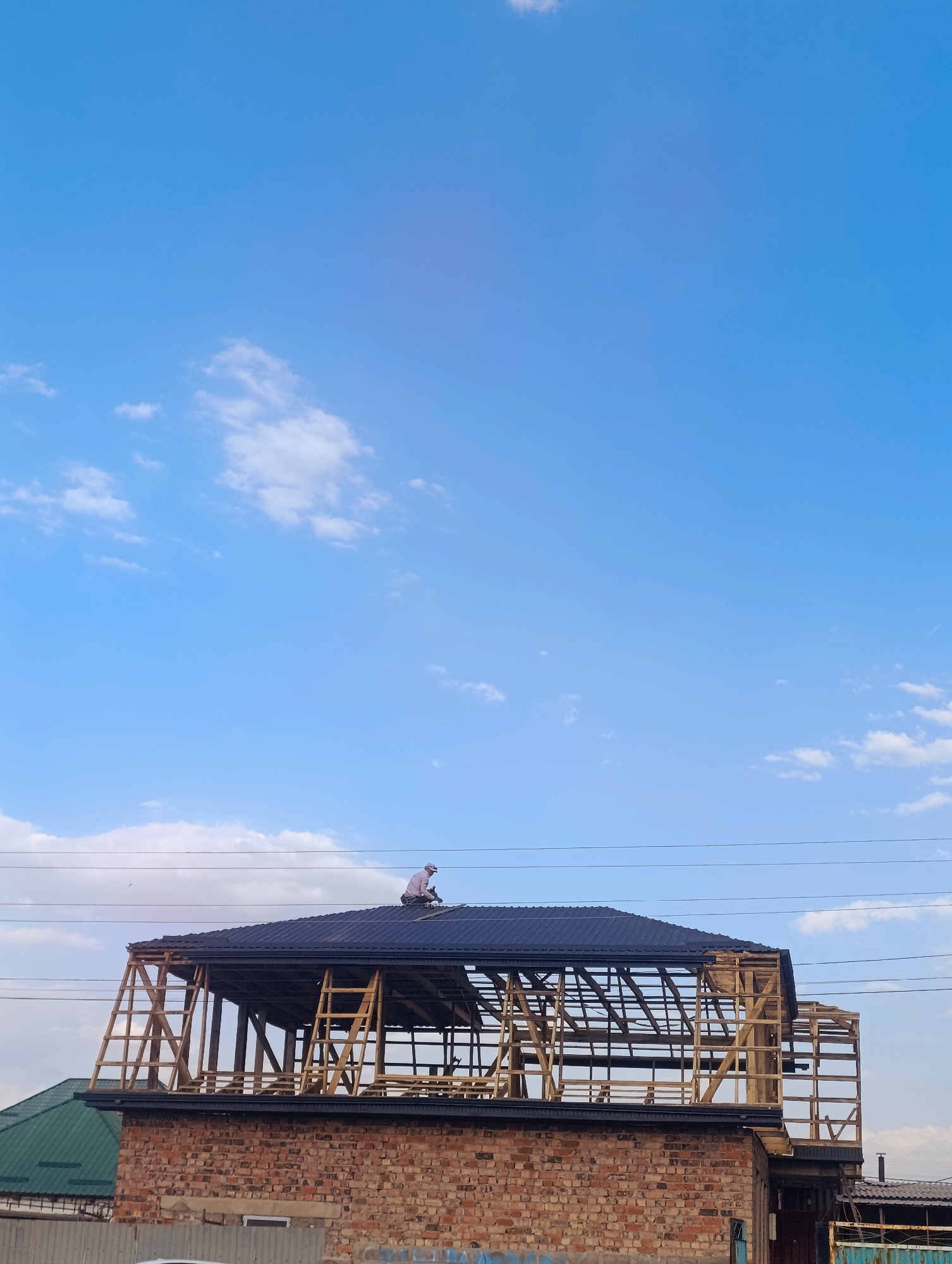 The image depicts a construction site in Naryn City, Kyrgyzstan, under a clear blue sky with scattered white clouds. The main focus is a partially constructed brick building with a dark metal roof. A person, wearing a light-colored shirt and a cap, is seen working on the roof, appearing to be using tools. Wooden scaffolding is erected around the upper section of the building, which is still open with exposed wooden beams. In the foreground, other structures and a metal fence are visible, suggesting a developed area within the city. Overhead power lines are also present in the background.